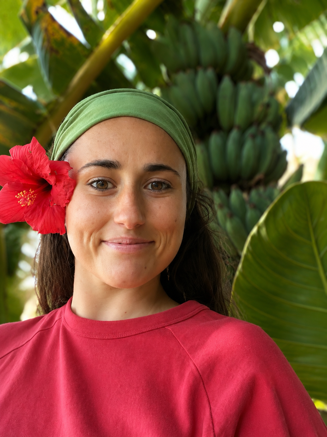 A woman with a pink flower in her hair, wearing a green headband and a red shirt, standing outdoors among lush green tropical plants and banana trees.