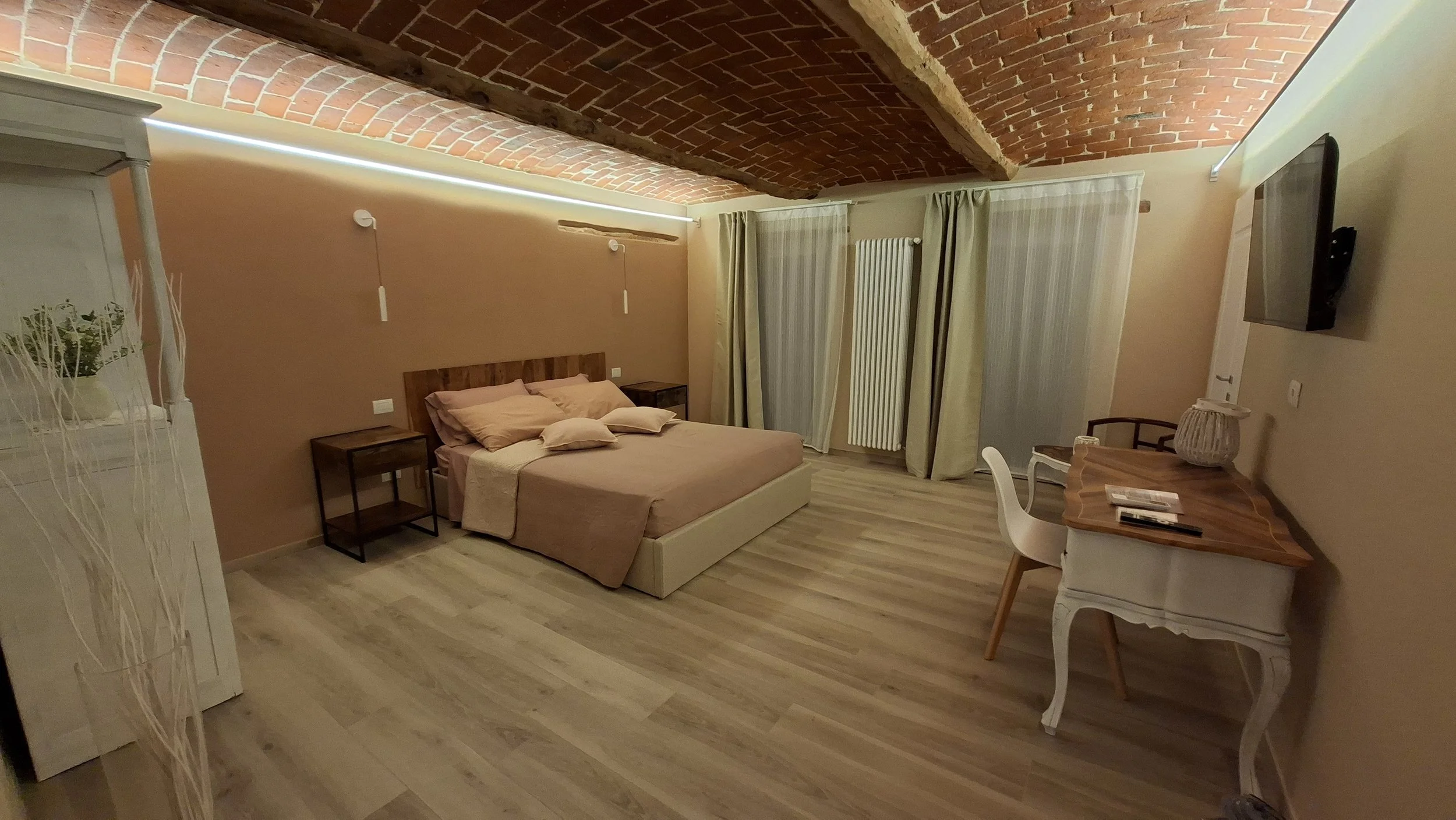 Una stanza da letto con soffitto in mattoni a vista, pareti color beige, letto con biancheria beige, due comodini in legno, tende chiare alle finestre, un radiatore bianco, un tavolo con una sedia bianca, una TV montata sulla parete.