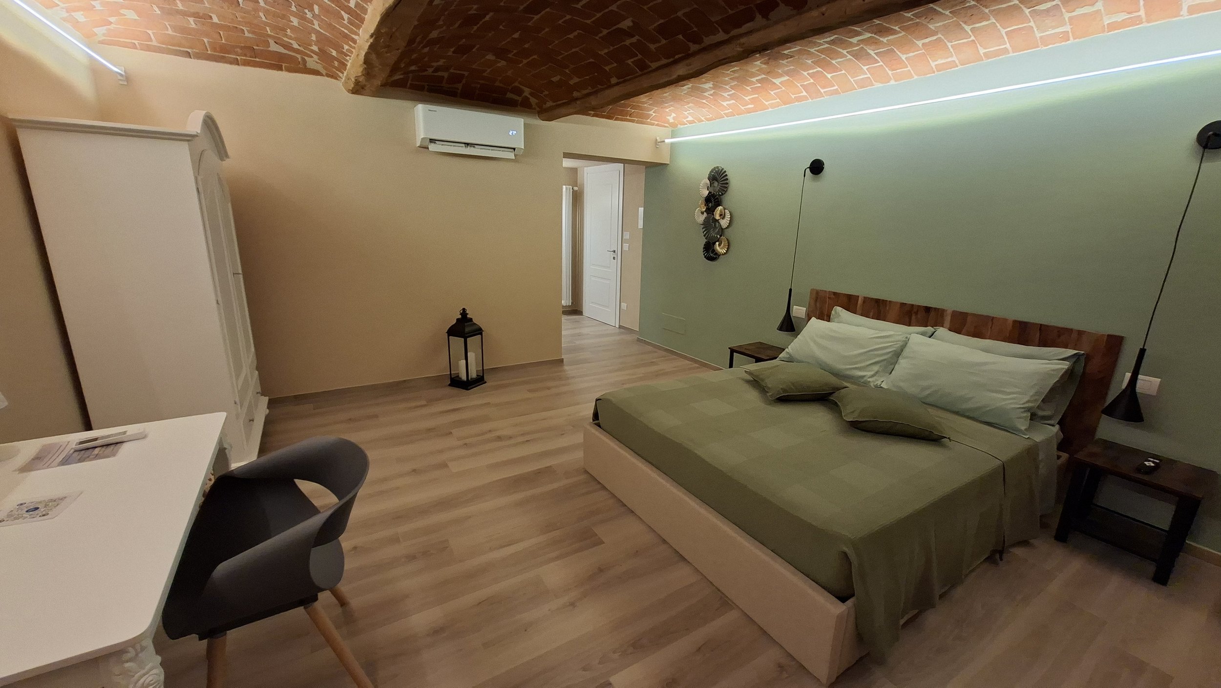 Camera da letto con letto matrimoniale, pareti di colore verde chiaro, soffitto a volta in mattoni, aria condizionata, decorazioni murali, lampade a sospensione nere, armadio bianco e tavolino.
