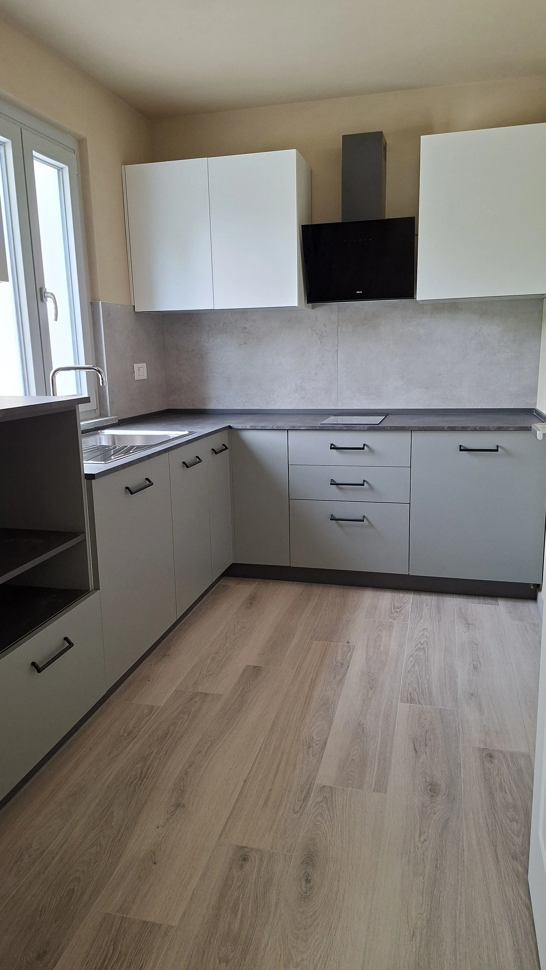 Cucina moderna con armadi bianchi e piano di lavoro in grigio, lavabo con rubinetto, cappa nera, pavimento in legno chiaro, finestra con vista esterna.