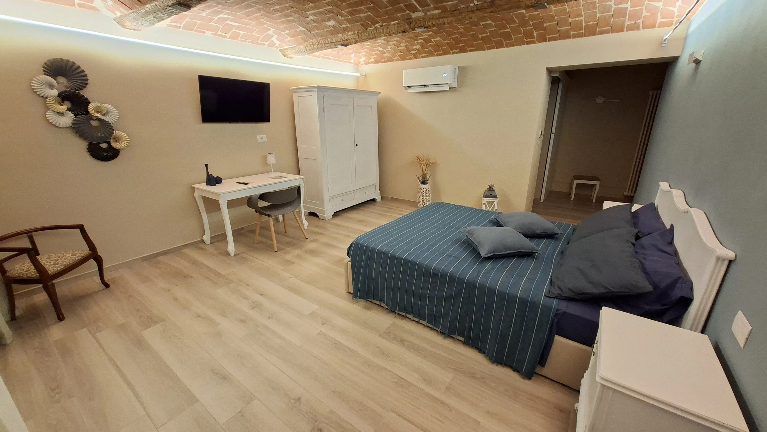 Camera da letto con pareti gialle, soffitto a volta in mattoni e pavimento in legno chiaro. Vi sono un letto con copriletto blu e cuscini grigi, un armadio bianco, una scrivania bianca con sedia grigia, una lampada da tavolo, una decorazione di ventagli sul muro, un televisore nero, un'unità di condizionamento e alcune piante e lanterne decorativi.