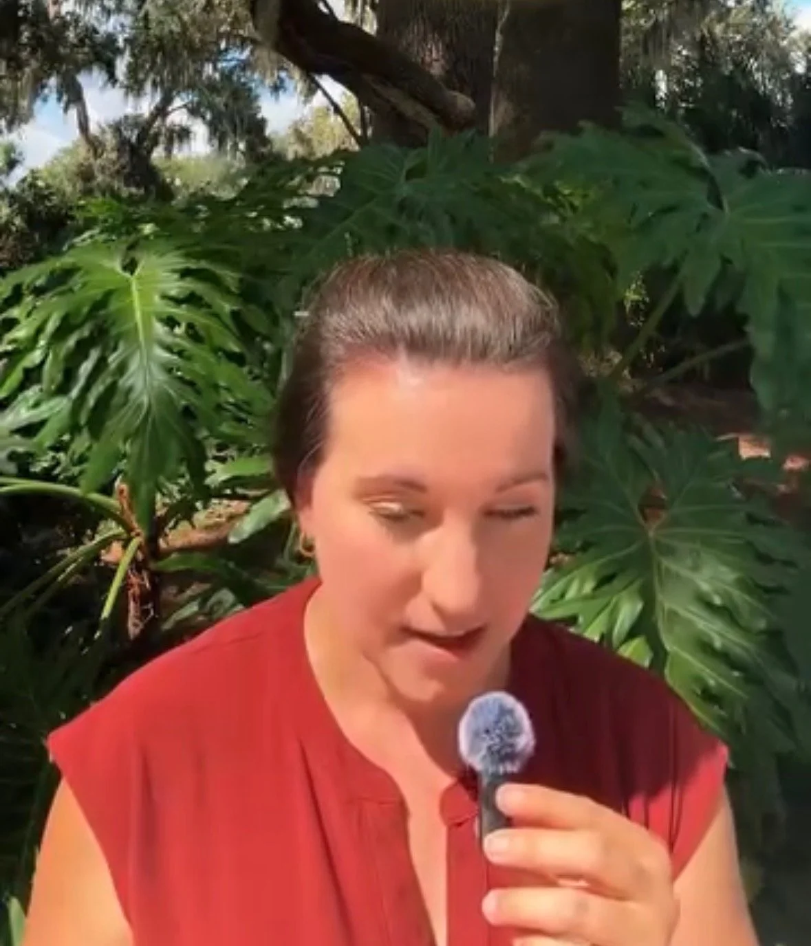 A woman holding a microphone with a fluffy windscreen recording a "Grounded is the New Sexy" podcast episode in nature, standing in front of large green leafy plants, outdoors with trees and cloudy sky in the background, wearing a red top.