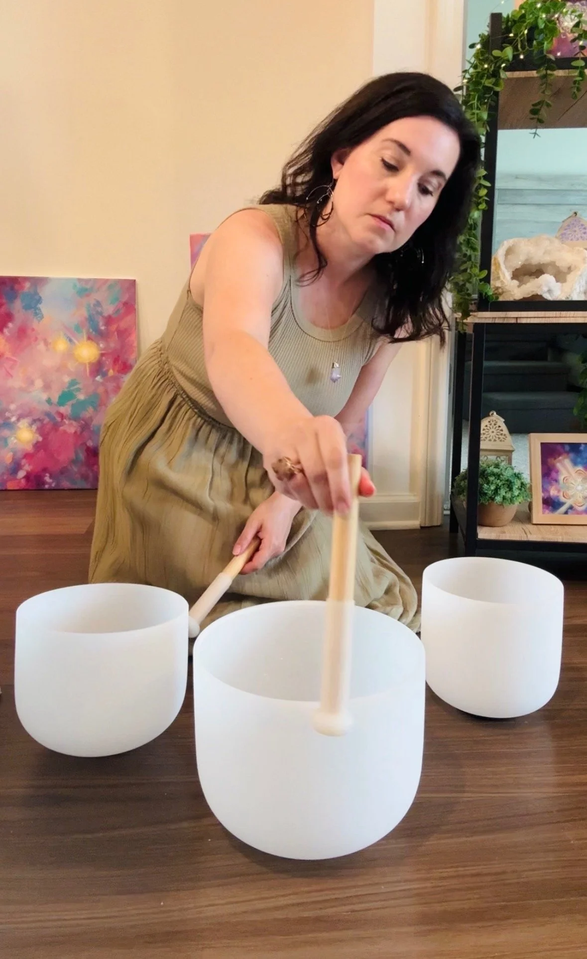 A woman is playing crystal singing bowls with mallets in a room decorated with energy artwork and plants.