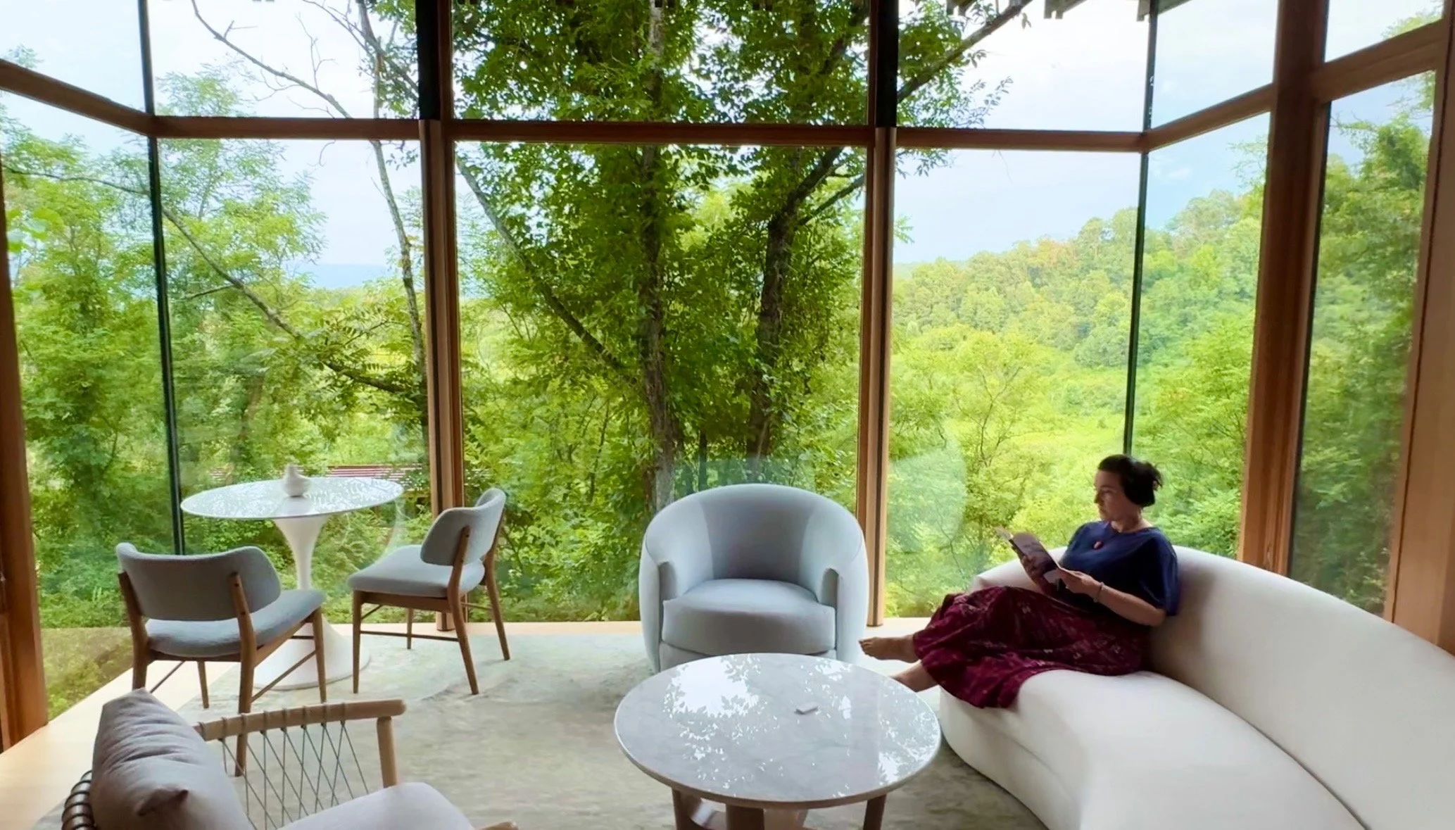 A woman sitting on a cream-colored couch, reading a book or tablet, in a room with floor-to-ceiling windows showcasing lush green trees outside.