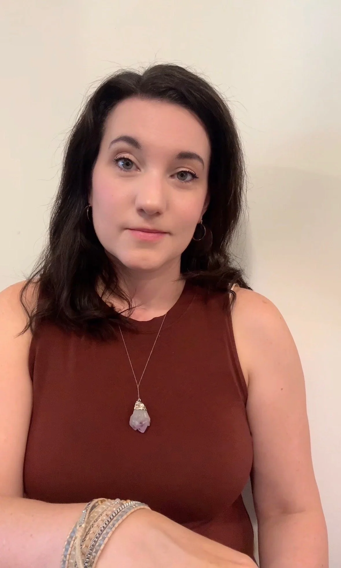 A woman with dark hair, wearing a sleeveless rust-colored top, a pinkish-purple pendant necklace, and multiple bracelets on her left wrist, posing against a plain off-white wall.