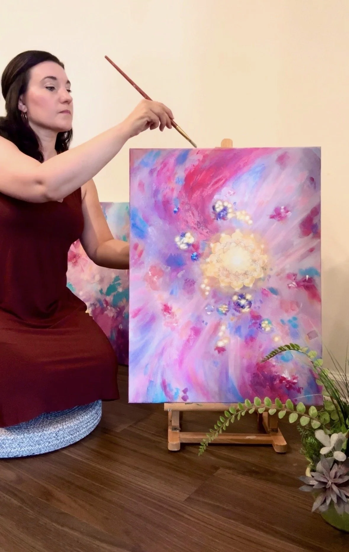 A woman in a maroon dress is painting on a colorful canvas with abstract swirls and bursts of pink, purple, blue, and white. She is kneeling on a wooden floor next to a green plant, with a plain beige wall in the background.