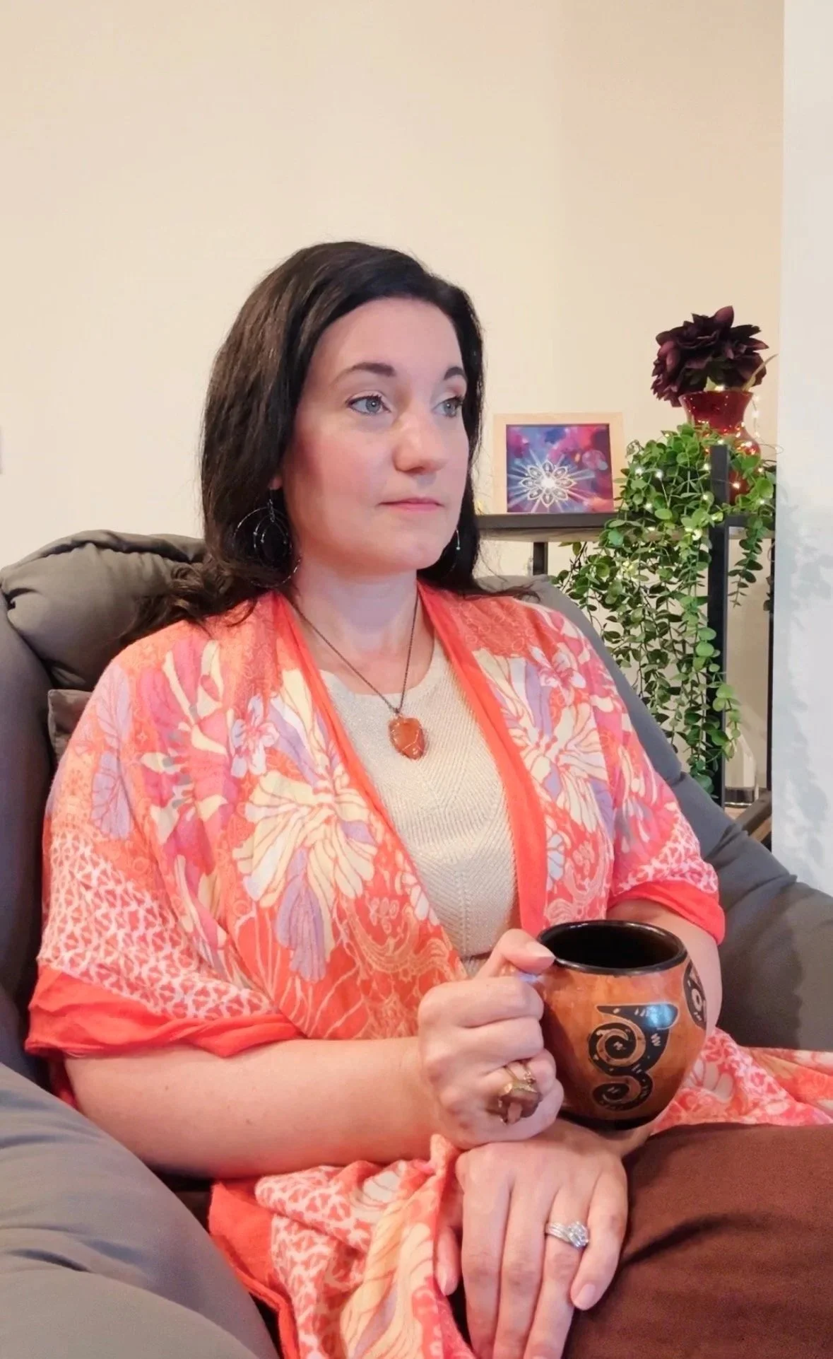 A woman with dark hair sitting on a couch, holding a decorated mug, wearing a beige top and a colorful orange and pink shawl, with plants and a framed artwork in the background.
