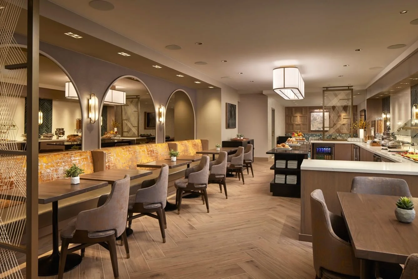 A spacious, well-lit hotel or restaurant dining area with a buffet setup, tables, and chairs, decorated with plants and modern lighting fixtures.