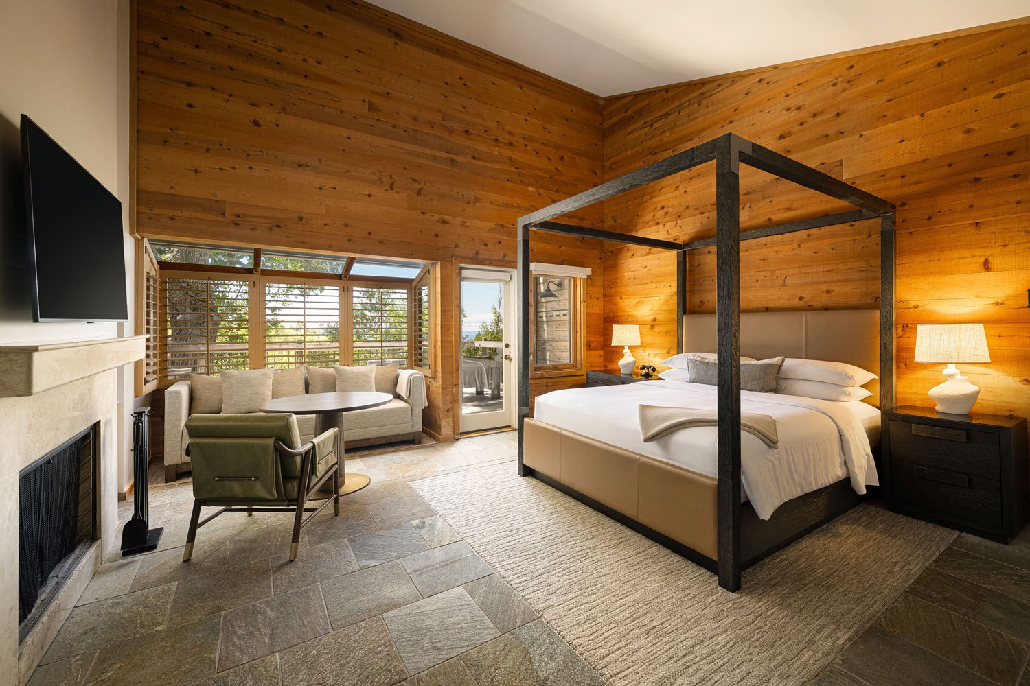 A bedroom with wooden walls and ceiling, featuring a large bed with a beige headboard and dark frame, two nightstands with lamps, a sitting area with a sofa and window, and a fireplace with a television above.