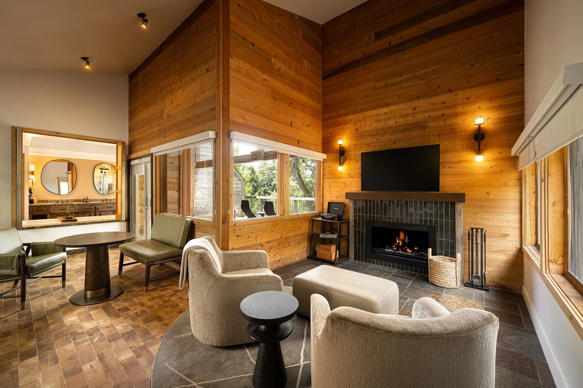 Living room with a cozy fireplace, wood-paneled walls, a television above the fireplace, large windows with blinds, a sitting area with armchairs and a round coffee table, and a view of an outdoor balcony with chairs.