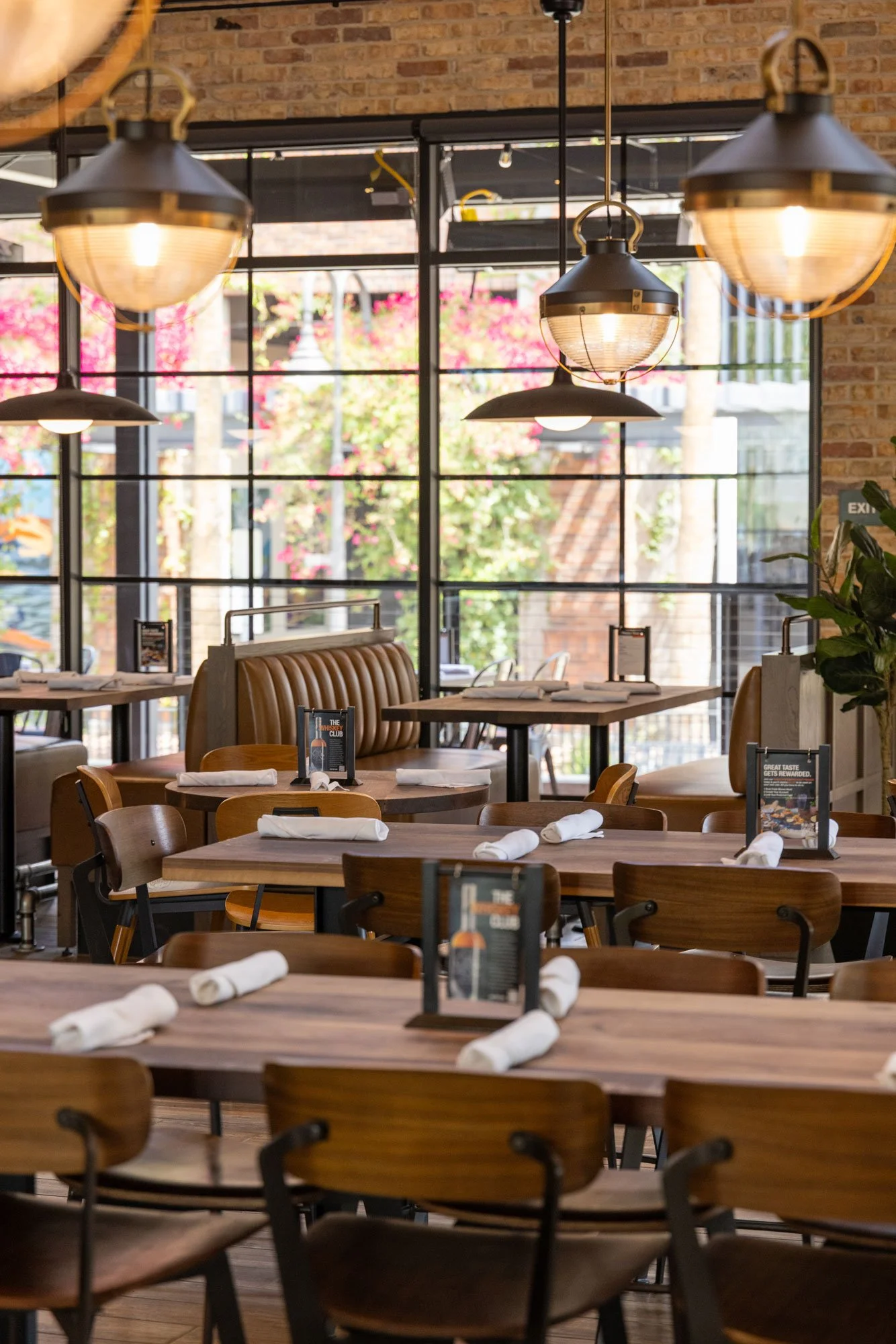 Interior of a restaurant with wooden tables, chairs, and booths, decorated with white napkins, and illuminated by hanging ceiling lights. Large windows reveal trees and buildings outside.