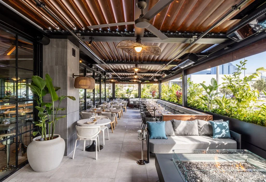 A bright, modern restaurant terrace with outdoor seating, greenery, and a mix of tables, chairs, and a sofa with cushions under a wooden ceiling.