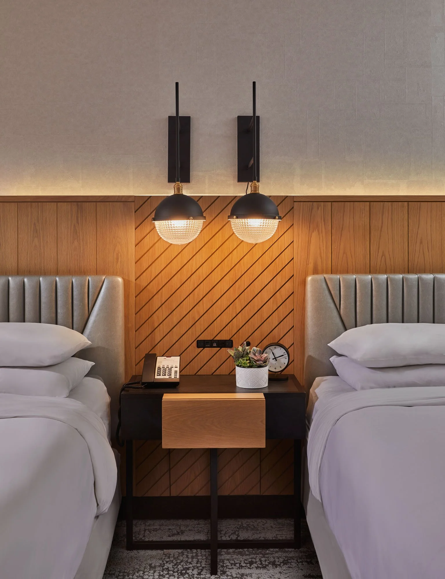 Two beds with white linens and padded headboards separated by a nightstand with a plant, clock, and cordless phone, in a room with a wooden accent wall and wall-mounted lamps.
