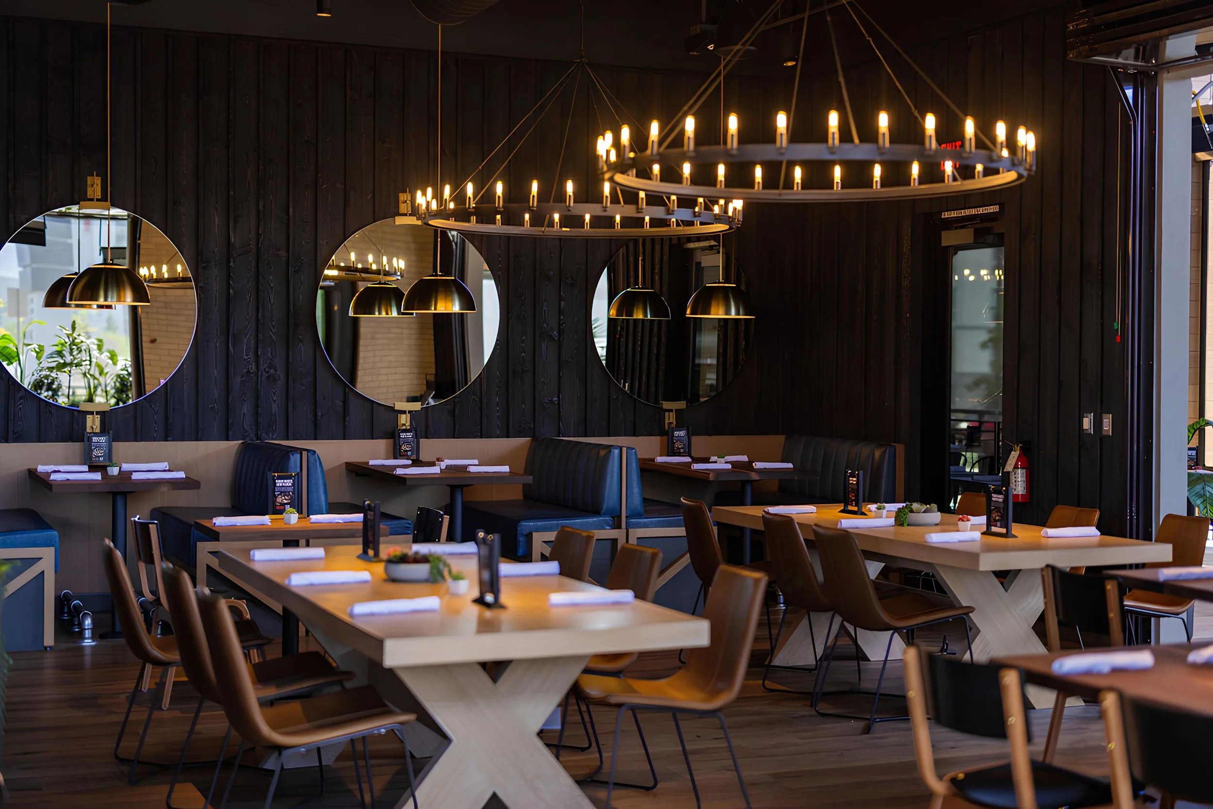 Interior of a modern restaurant with wooden furniture and circular chandeliers with candle-like bulbs, black paneled walls, and large mirrors reflecting the space.