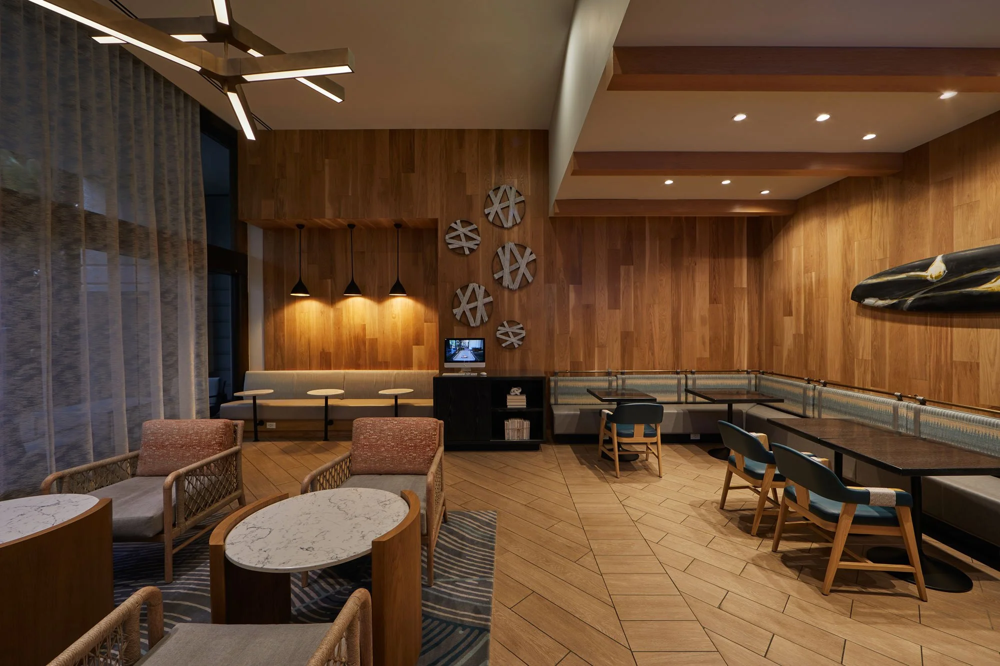 Interior of a modern hotel lobby or lounge area with wood-paneled walls, hanging pendant lights, decorative wall art, and a variety of seating options including armchairs and sofas, with a marble-topped coffee table and a striped rug.