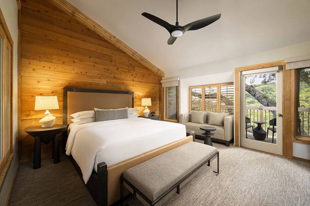 Modern bedroom with a wooden accent wall, a large bed with white bedding, two bedside lamps, a sofa with cushions, a bench at the foot of the bed, and a balcony door with a view of trees outside.