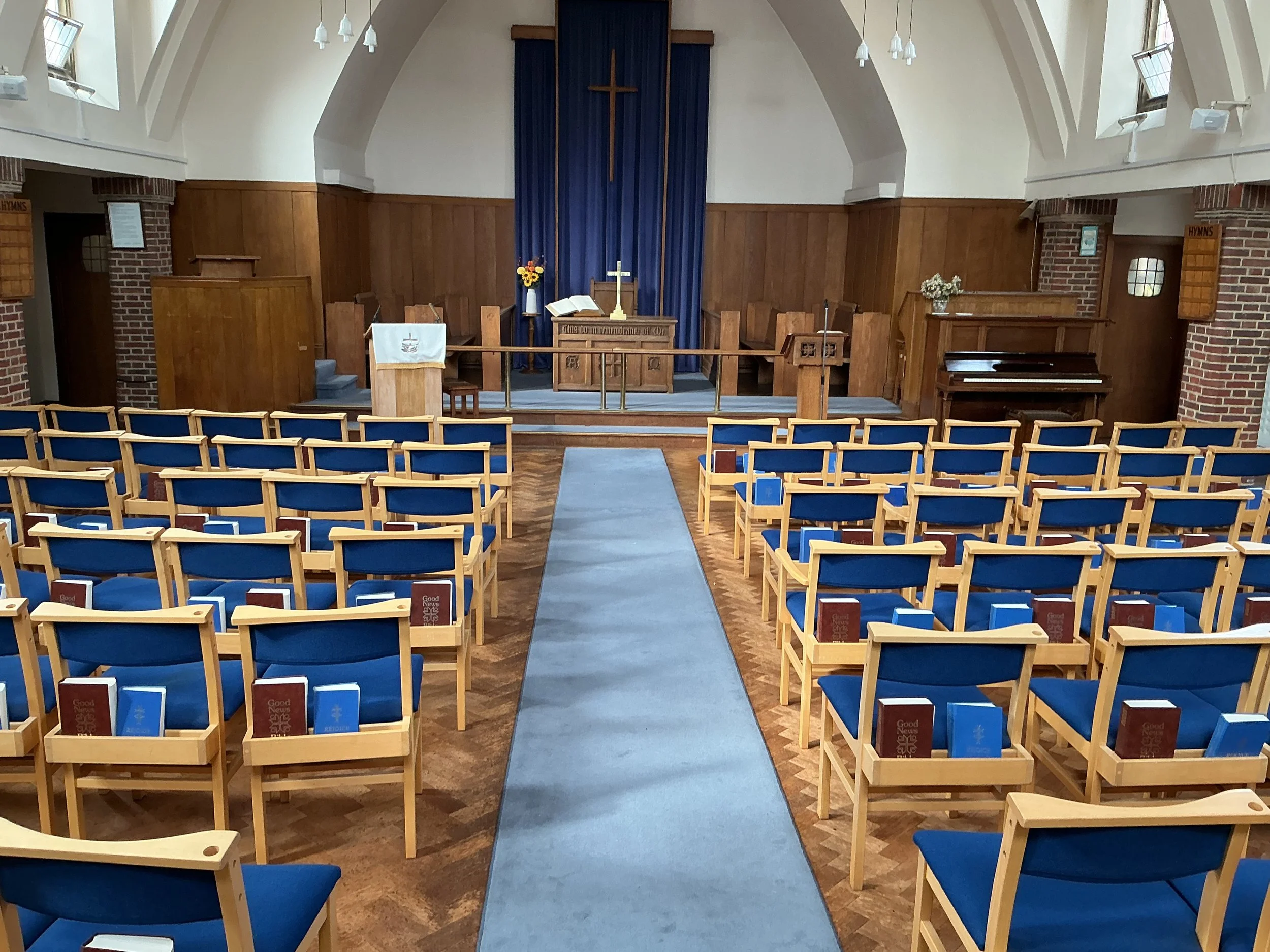 Interior of a church with wooden pews, a blue carpet aisle, altar at the front with a cross, flowers, and an open Bible, brick walls, and windows providing natural light.