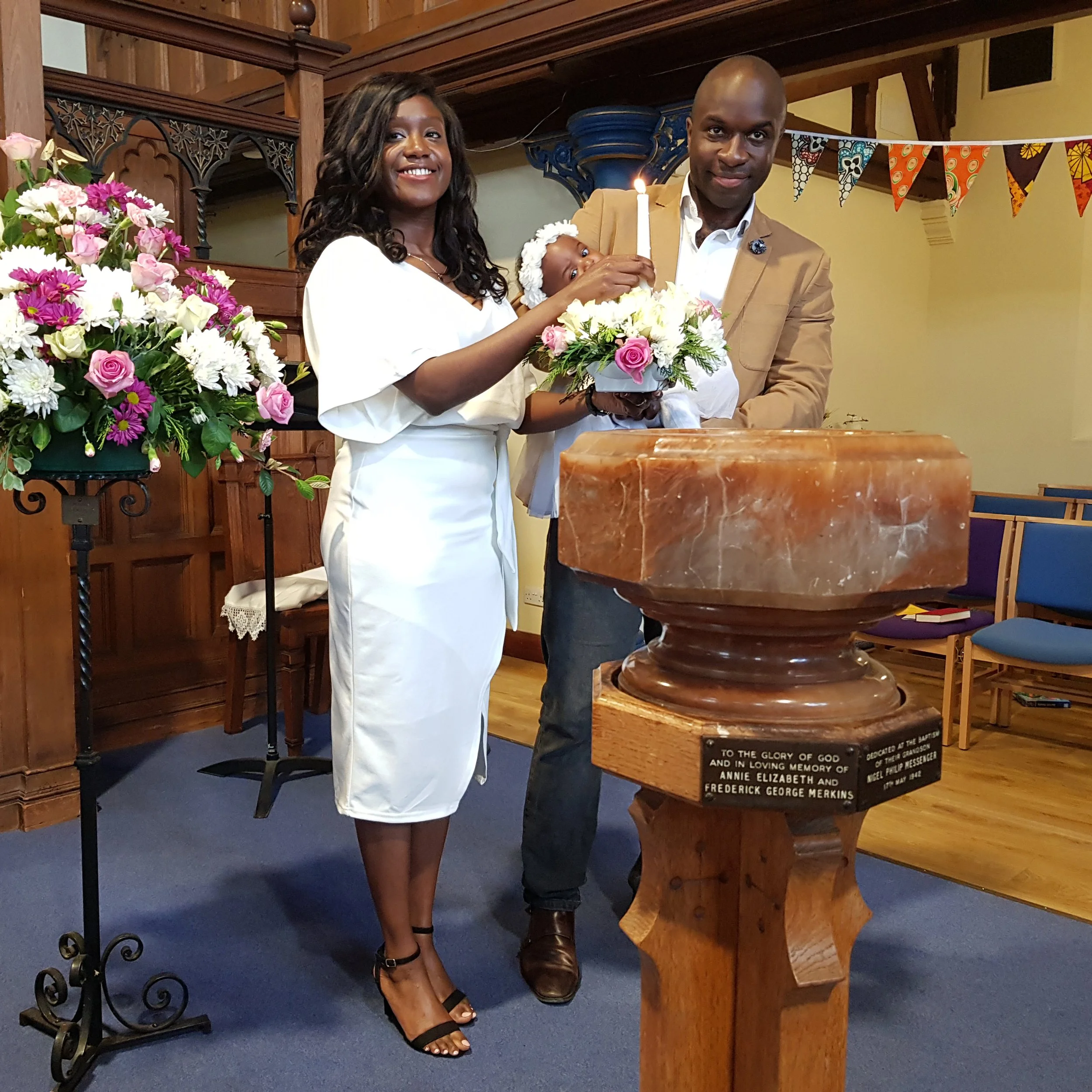 A woman and a man are standing behind a baptismal font, holding a baby with a lit candle. The woman is smiling and wearing a white dress, while the man is wearing a tan jacket and the woman is wearing black high heels. The scene appears to be in a church, decorated with flowers, wood paneling, and colorful pennant banners.