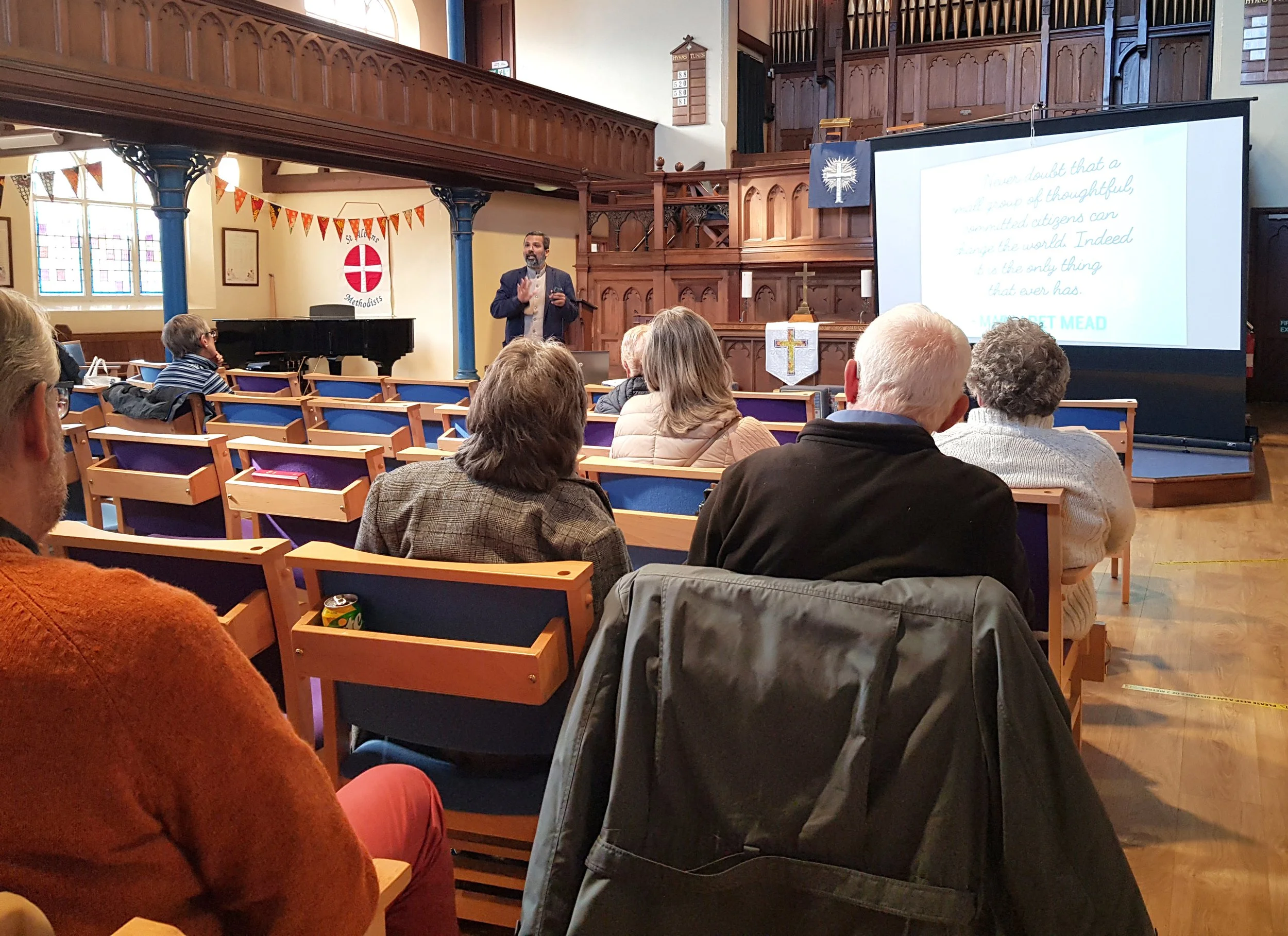 A man in a suit speaking to an audience inside a church, with a large screen displaying a quote by Margaret Mead. The church has wooden details, stained glass windows, and a banner with a cross hanging on the wall.