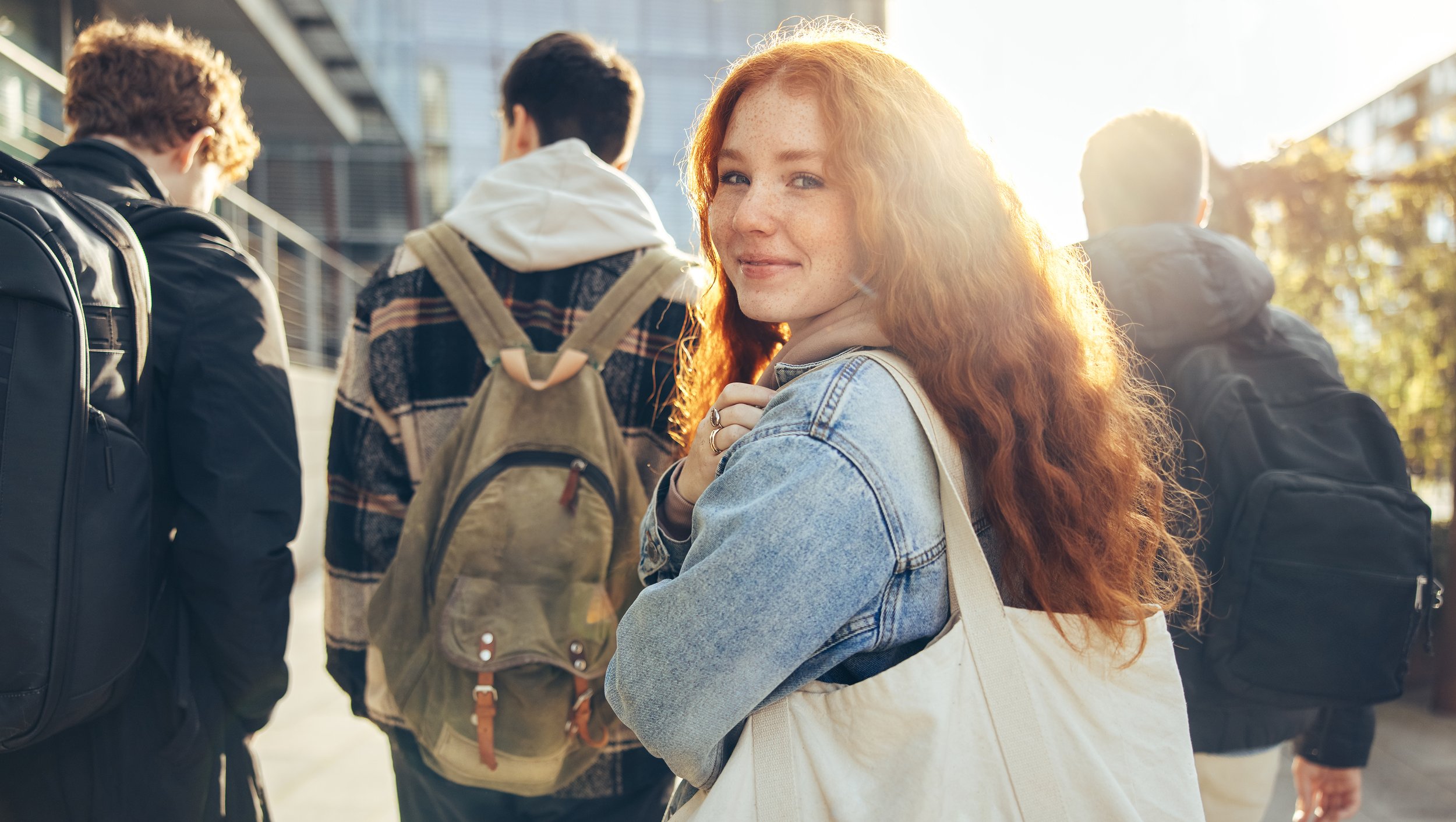A red-haired woman with freckles smiling at the camera, standing among a group of people with backpacks, outdoors in sunlight, in an urban setting.