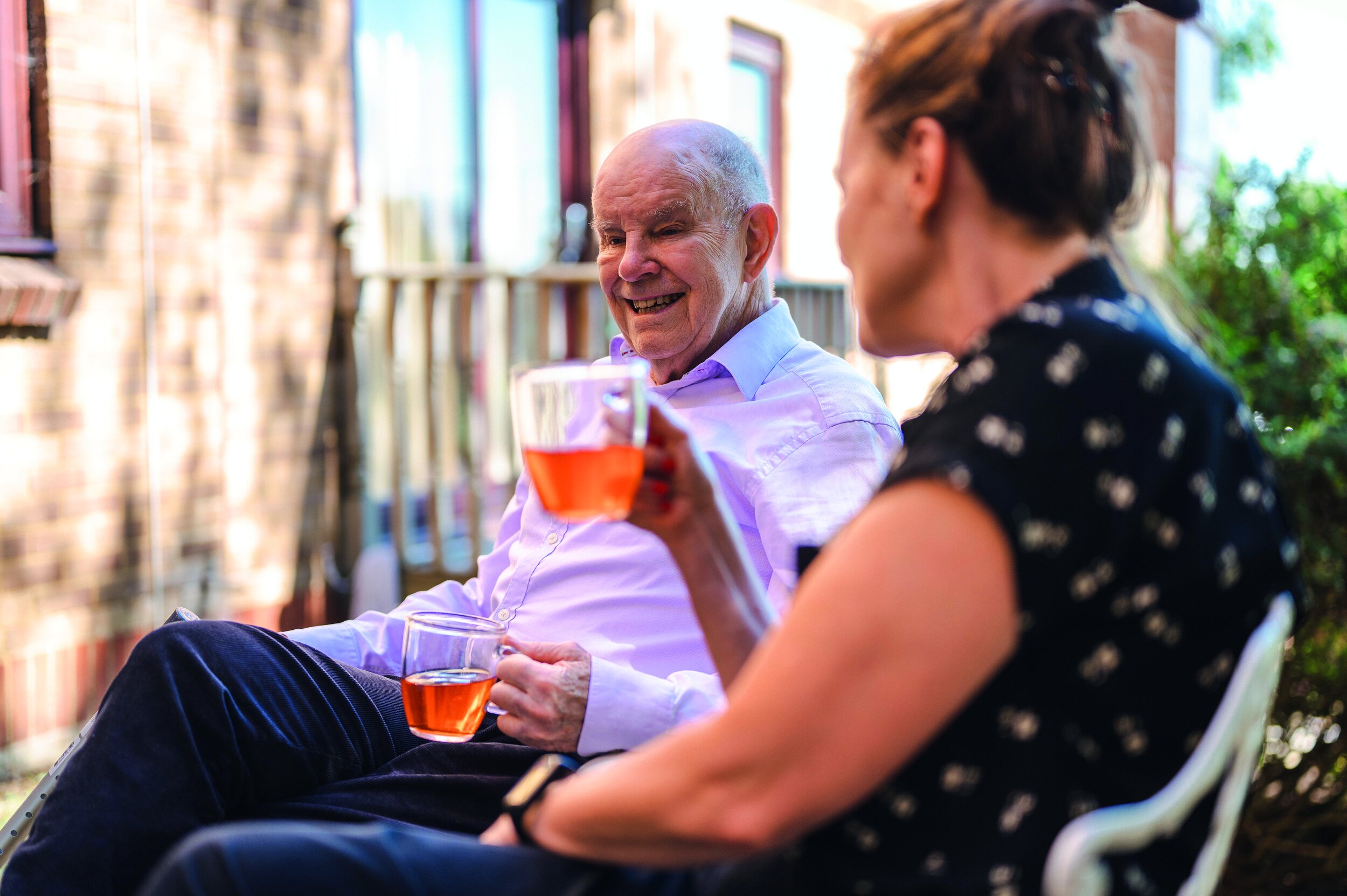 Two people sitting outdoors, holding glasses of pink drinks, engaging in conversation; one is an elderly man with a white shirt, and the other is a woman with dark hair in a bun and a dark patterned shirt, with a brick building and greenery in the background.
