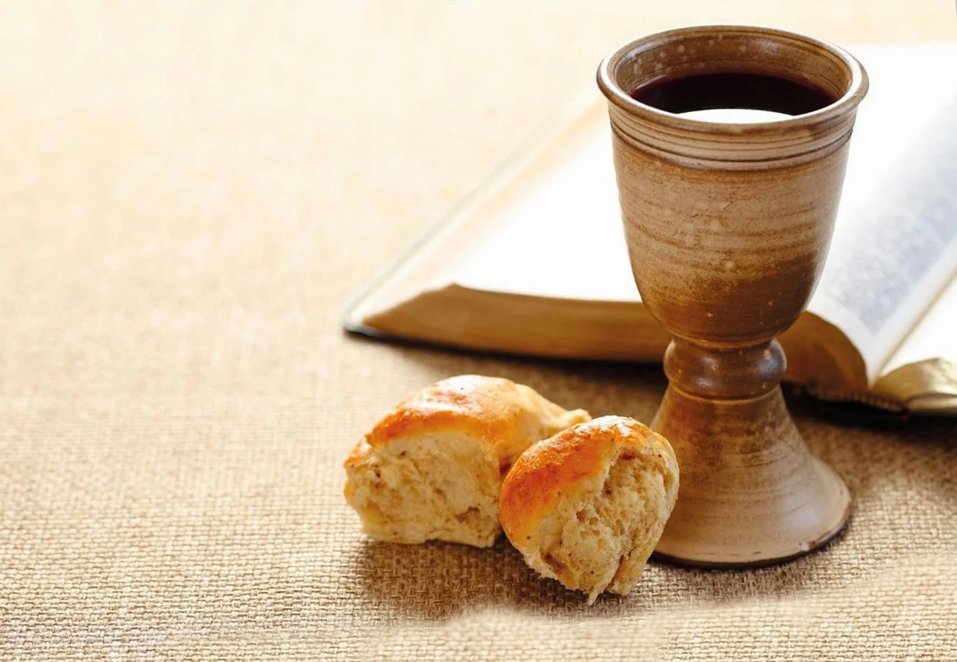 A wooden chalice filled with red wine, two scones, and an open book on a textured beige surface.