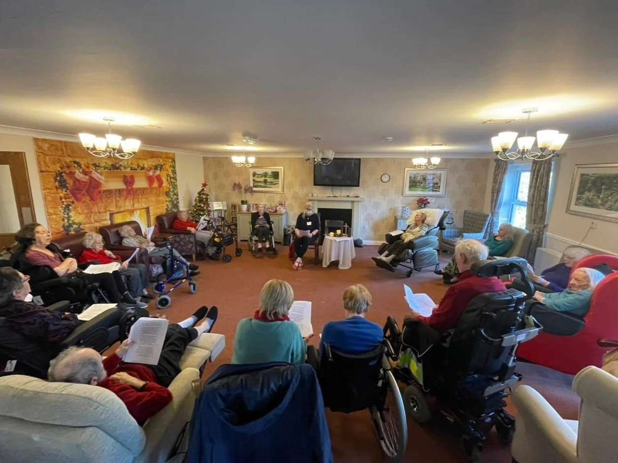 Group of elderly people sitting in a circle in a living room, some in wheelchairs, participating in a group activity or meeting.