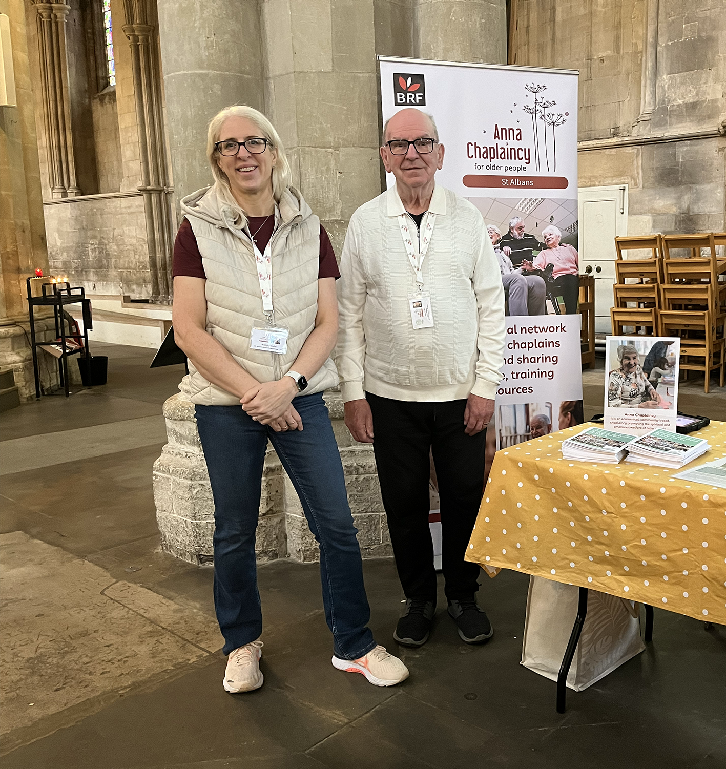 Two people standing in front of a display booth with a banner about Anna Chaplaíncy for older people. The woman on the left is wearing a beige vest over a maroon shirt, glasses, and jeans, and the man on the right is wearing a white sweater and black pants. There are informational brochures on a table covered with a yellow polka dot cloth.