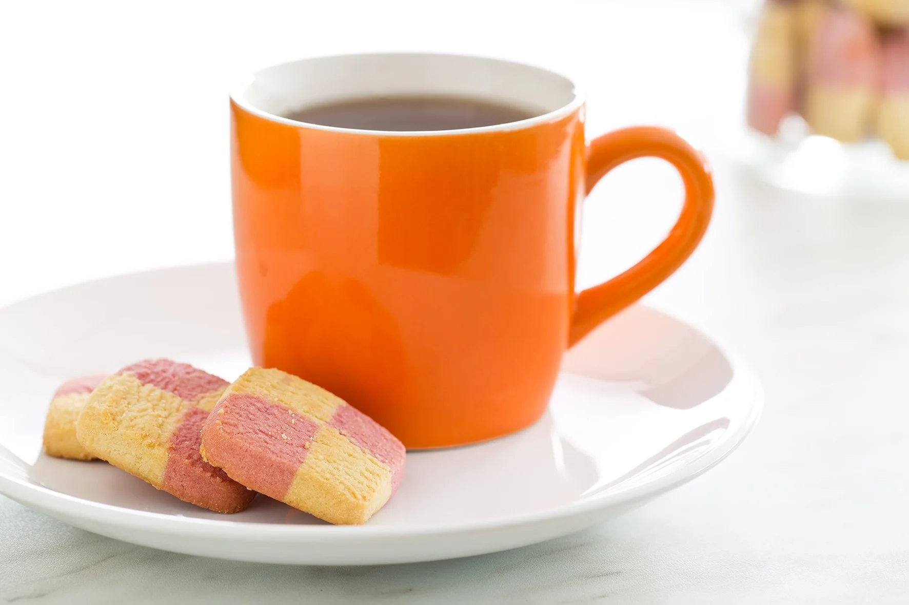 Orange mug filled with coffee or tea, on a white plate with two striped pink and yellow cookies, on a light-colored surface.