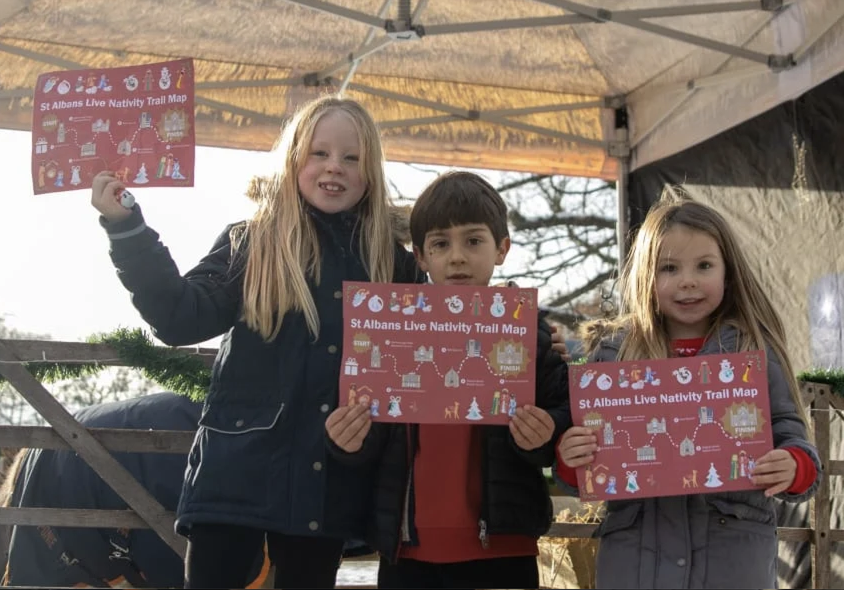 Three children holding red trail maps titled "St Albans Live Nativity Trail Map" at an outdoor event with a tent and trees in the background.