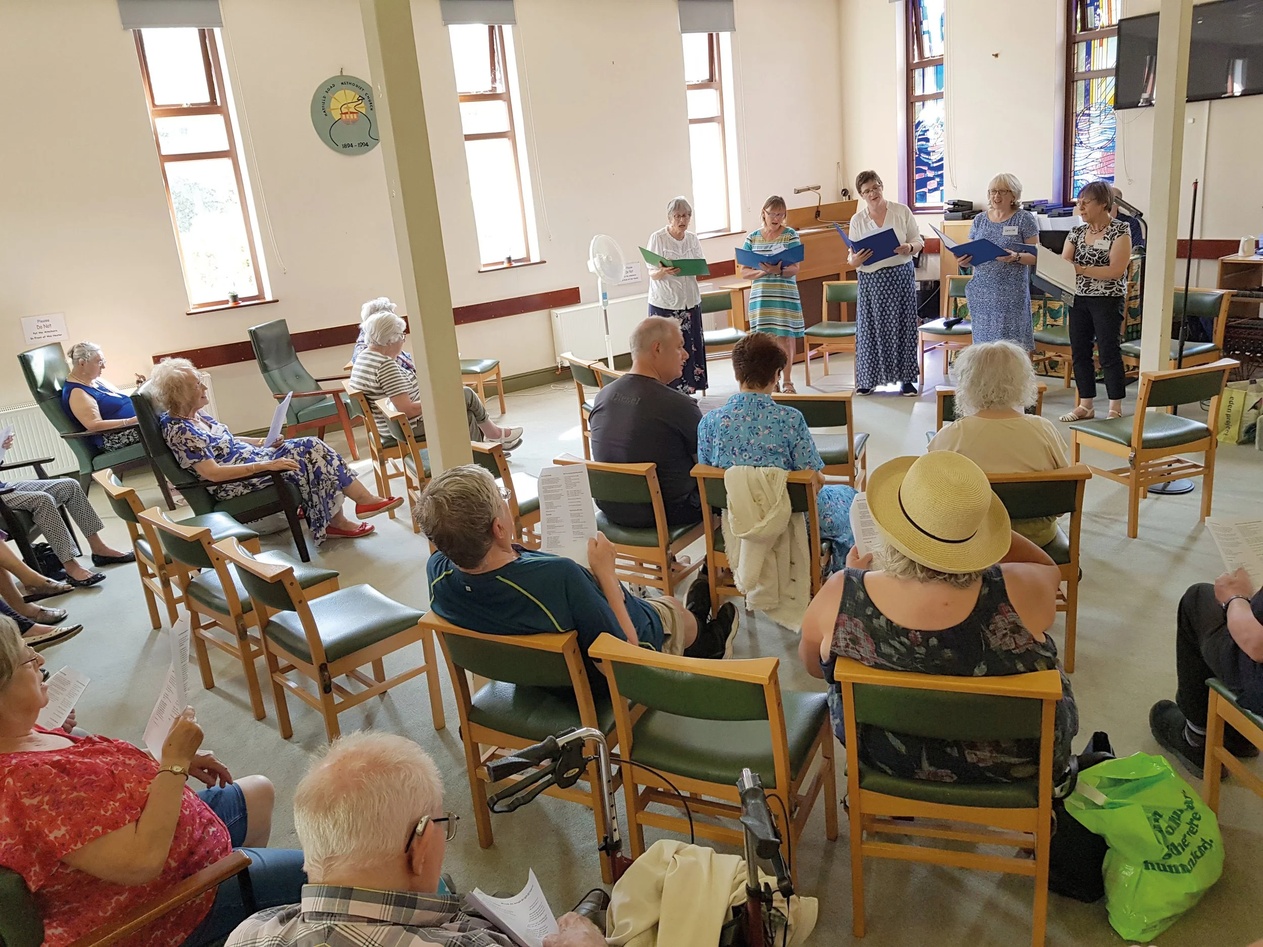 A group of elderly women and men singing in a church or community center, holding songbooks, with stained glass windows in the background, and seated on chairs facing the choir.