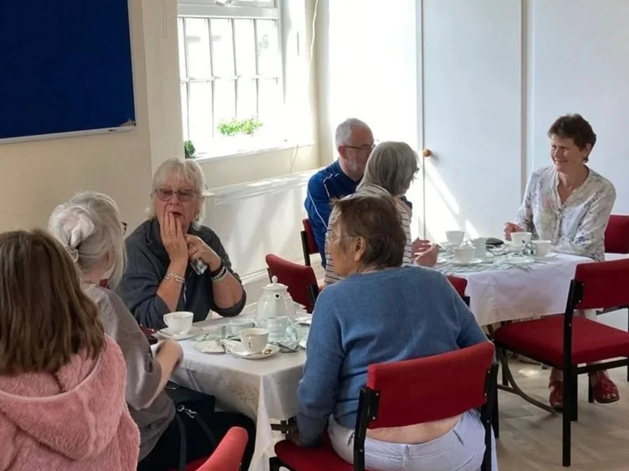 Group of older adults sitting around a table, having tea or coffee, engaging in conversation, in a well-lit room with a window and a blue chalkboard or notice board.
