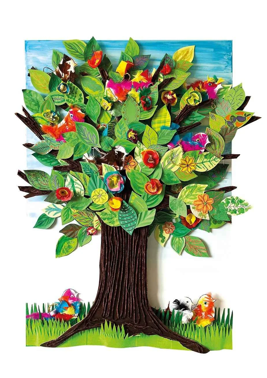 Colorful paper craft of a tree with various birds and flowers, set against a painted blue sky background. The tree has a brown trunk and green leaves, with some birds perched on or flying around it, and a grassy ground at the bottom.