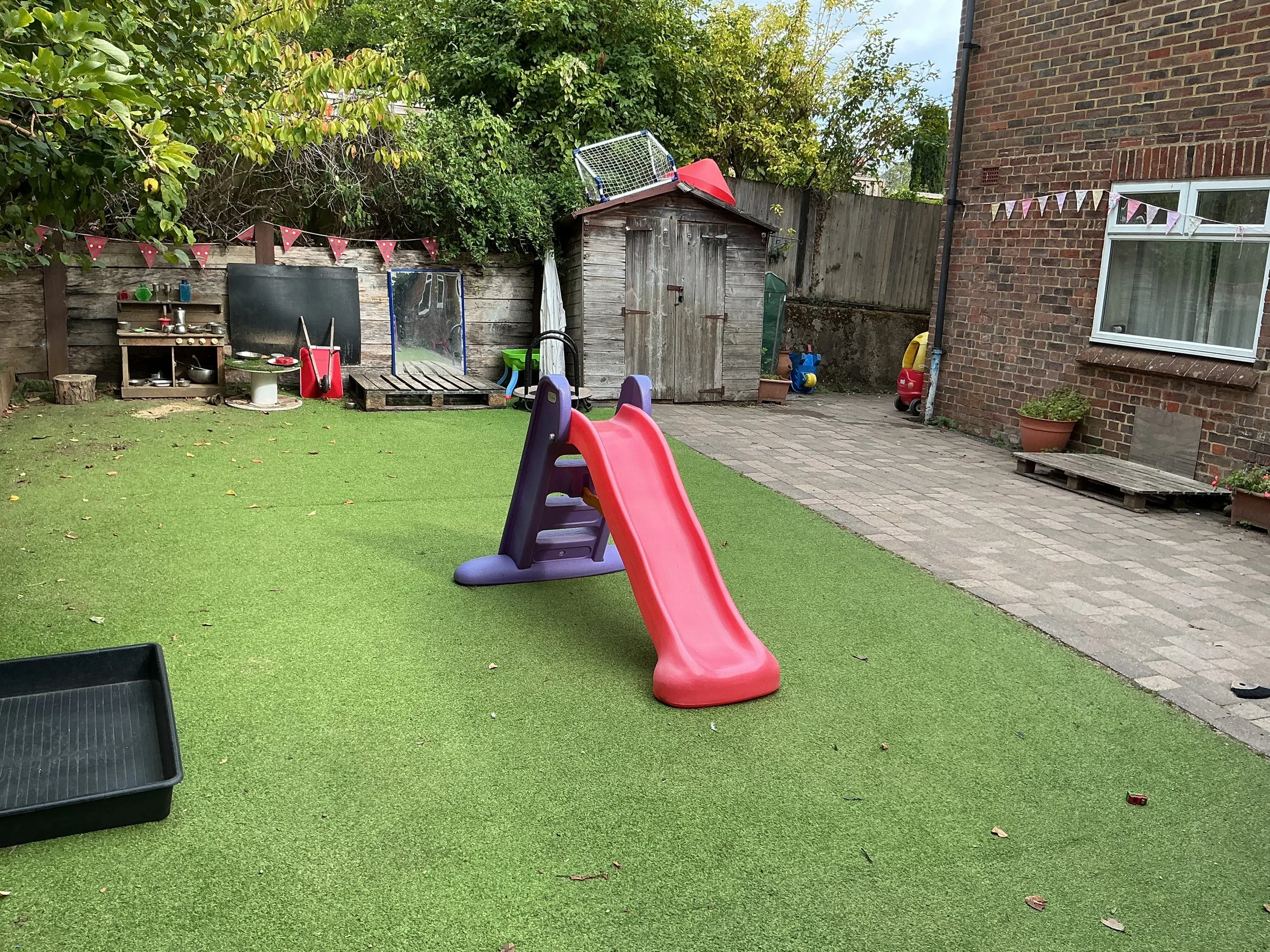 Backyard with a red slide, small wooden shed, garden, and various children's toys on a patio and grass area.