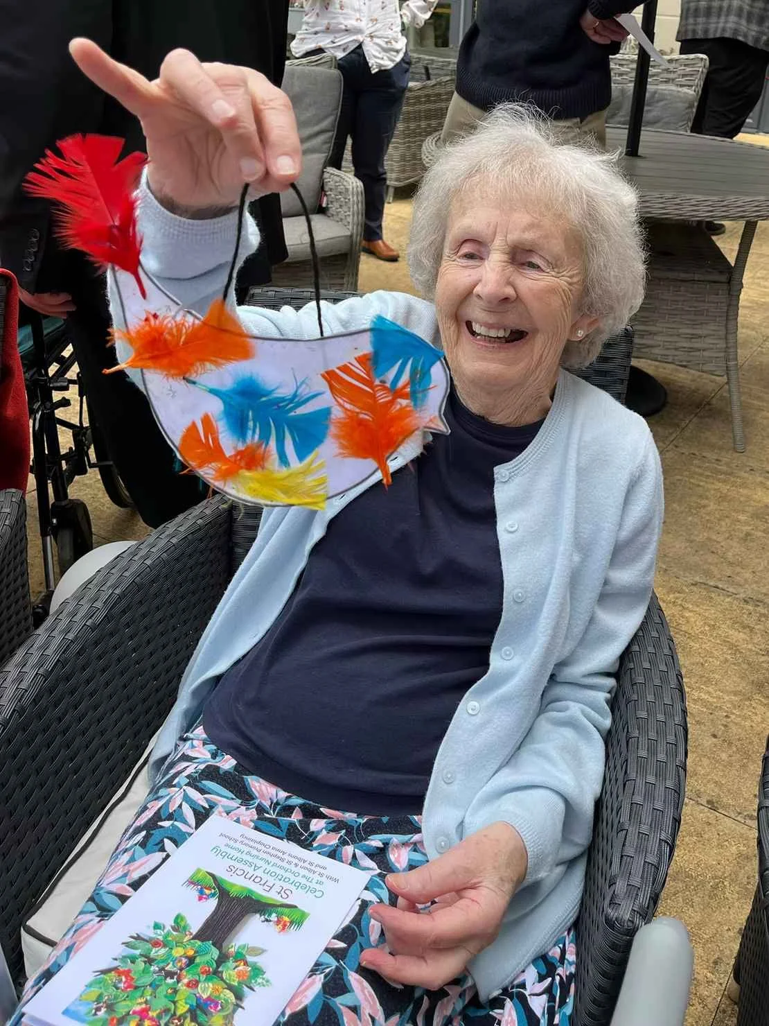 An elderly woman sitting in a wheelchair, smiling, as someone holds up a colorful feathered face mask in front of her. The woman has white hair and is dressed in a light blue cardigan and patterned pants. There are other people in the background at an outdoor gathering.