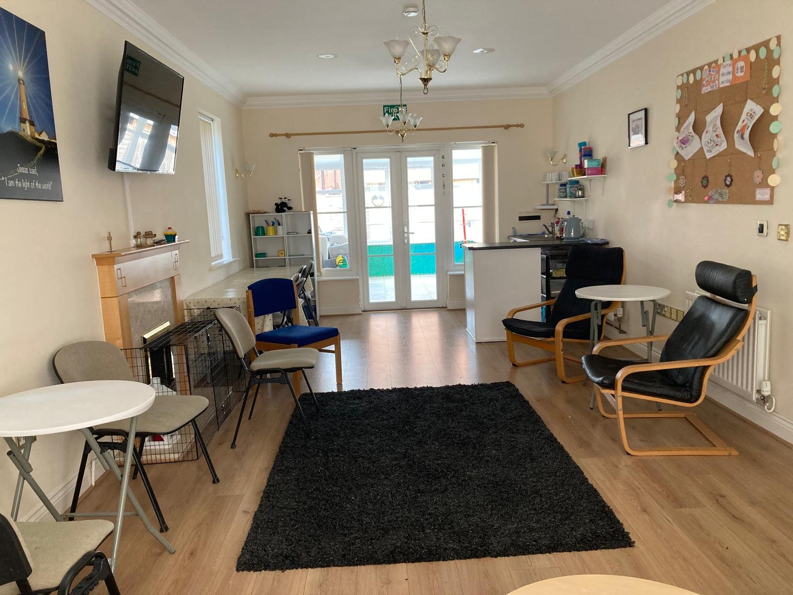 Living room with multiple chairs, small tables, a black rug, and a kitchenette area with white cabinets and shelves. A large window and glass doors lead to a balcony, with a chimney and artwork on the walls.