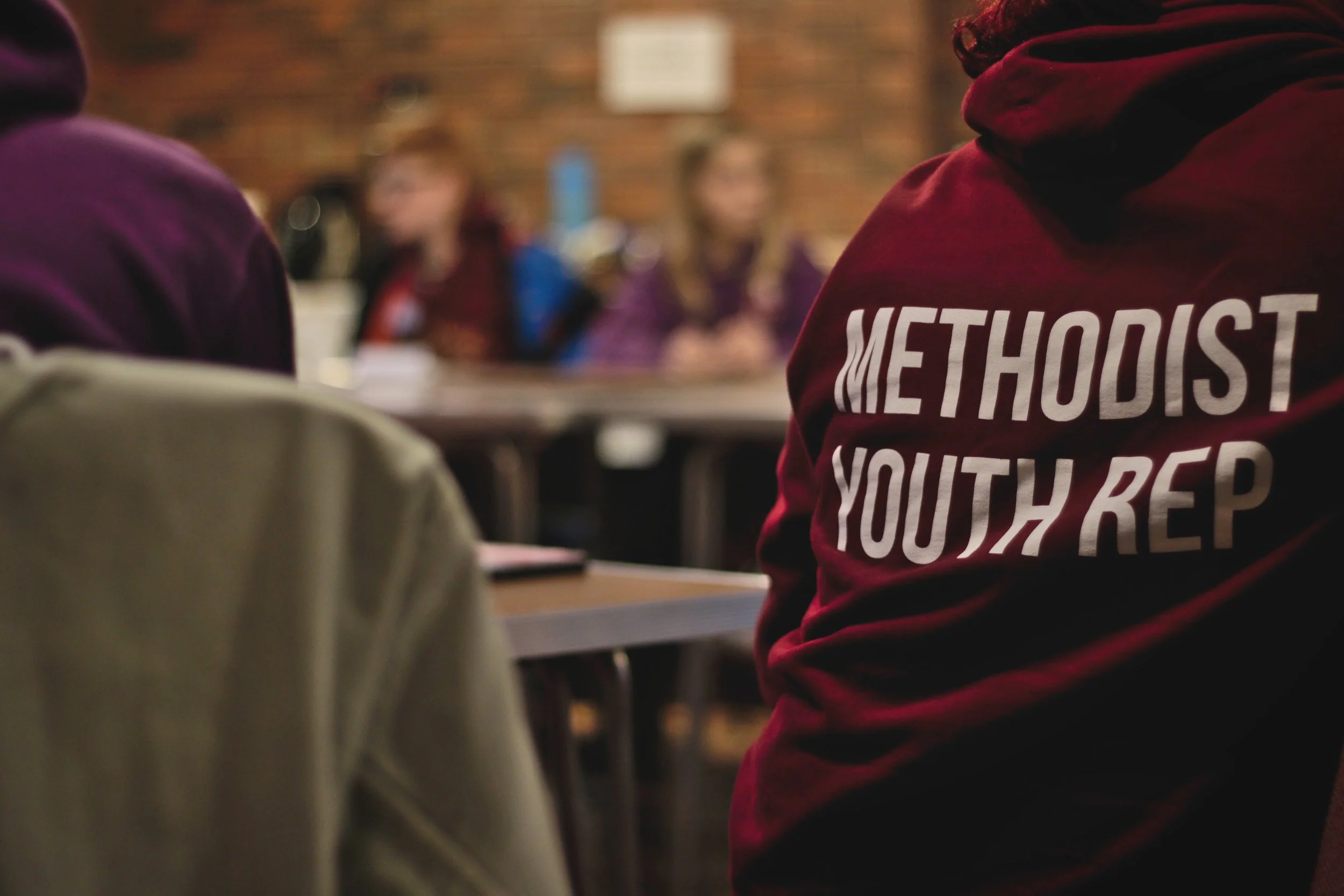 A person wearing a red hoodie with white text on the back, sitting in a room with exposed brick walls and other people in the background, some blurred.