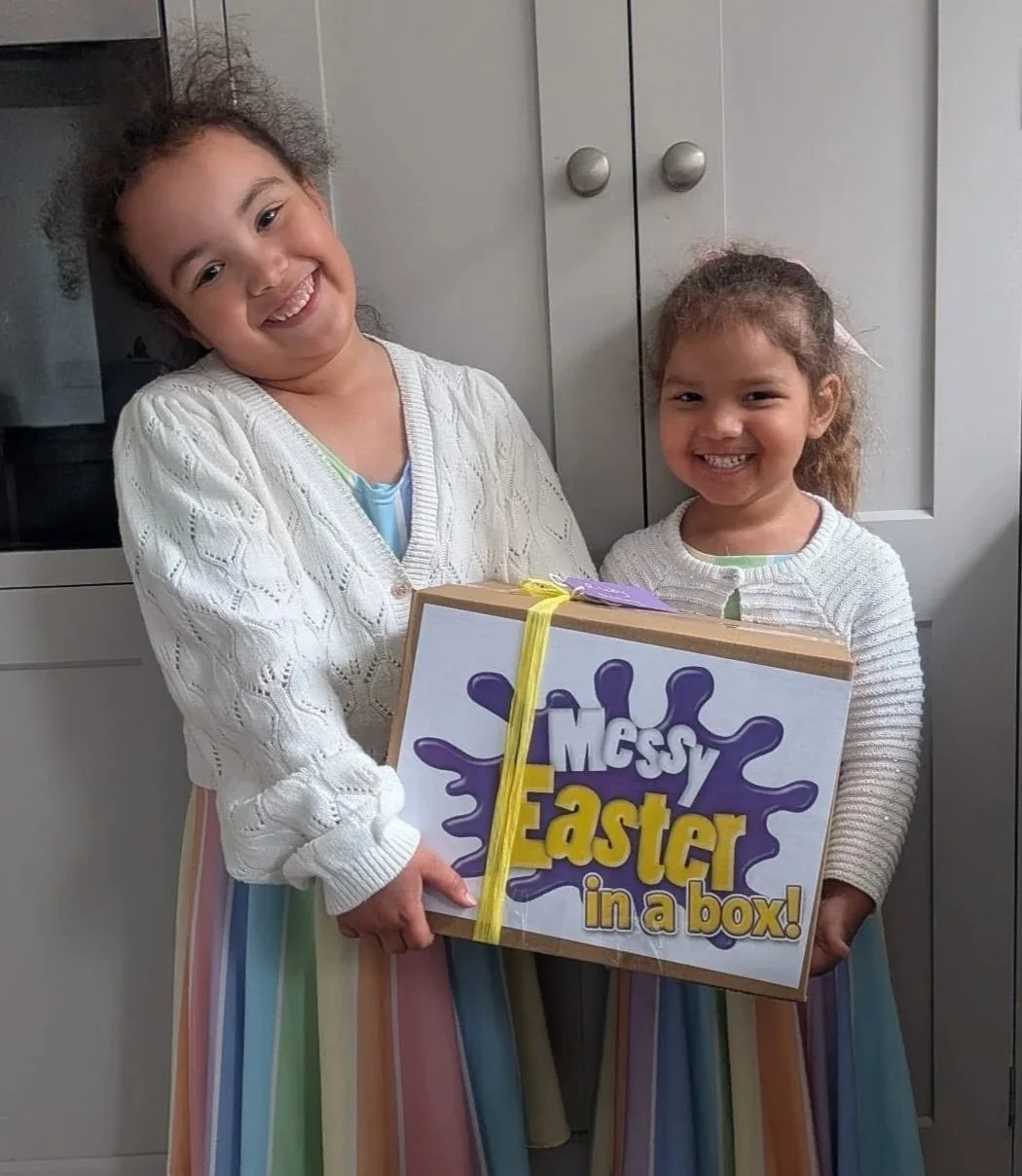 Two young girls smiling and holding an Easter gift box labeled 'Messy Easter in a box!' in a kitchen.