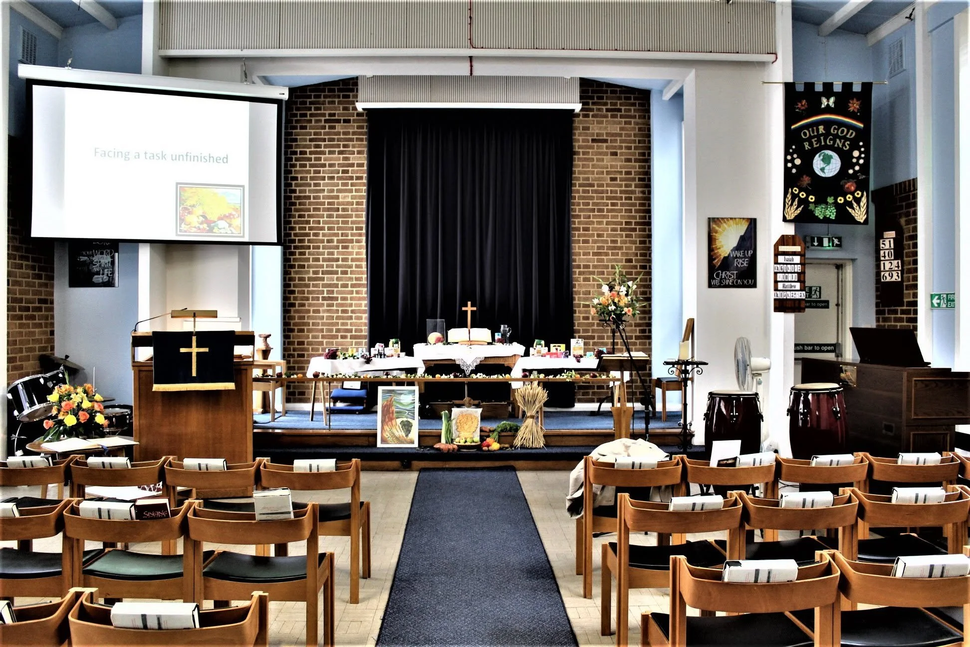 Interior of a church with chairs arranged facing the altar. The altar has flowers and religious items, with a cross at the center. There is a large projection screen on the left and banners on the right, with one banner reading 'Our God Reigns.' The background shows brick walls and a black curtain behind the altar.