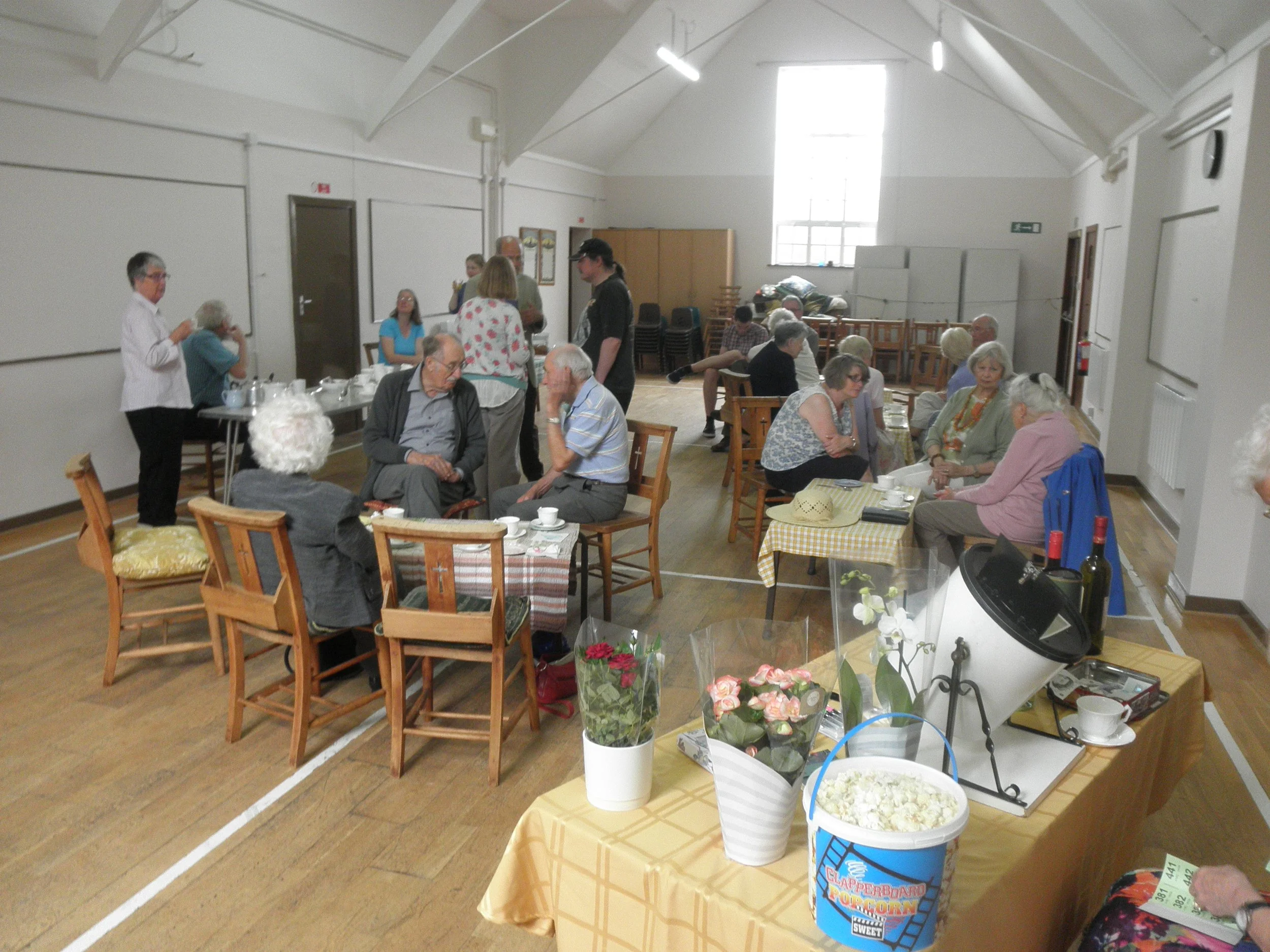 People seated at tables in a large room, engaging in conversation, with a table of food and flowers in the foreground.