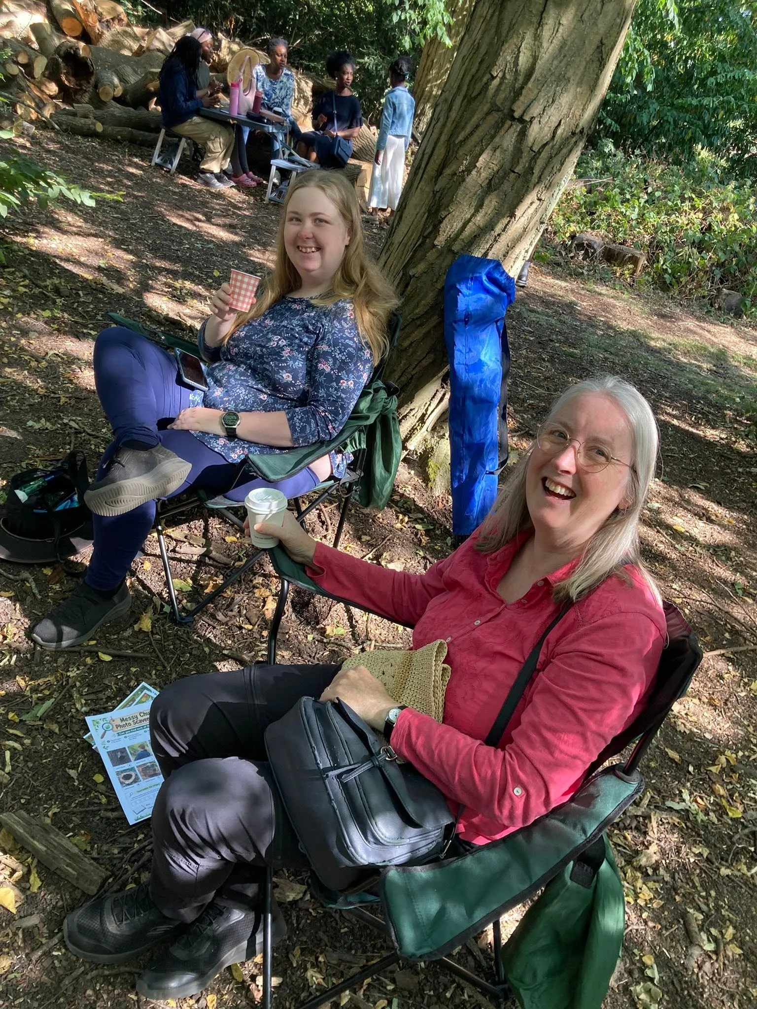 People sitting on chairs outdoors in a wooded area, smiling, with one woman holding a cup, and a group in the background sitting and standing near a large tree.