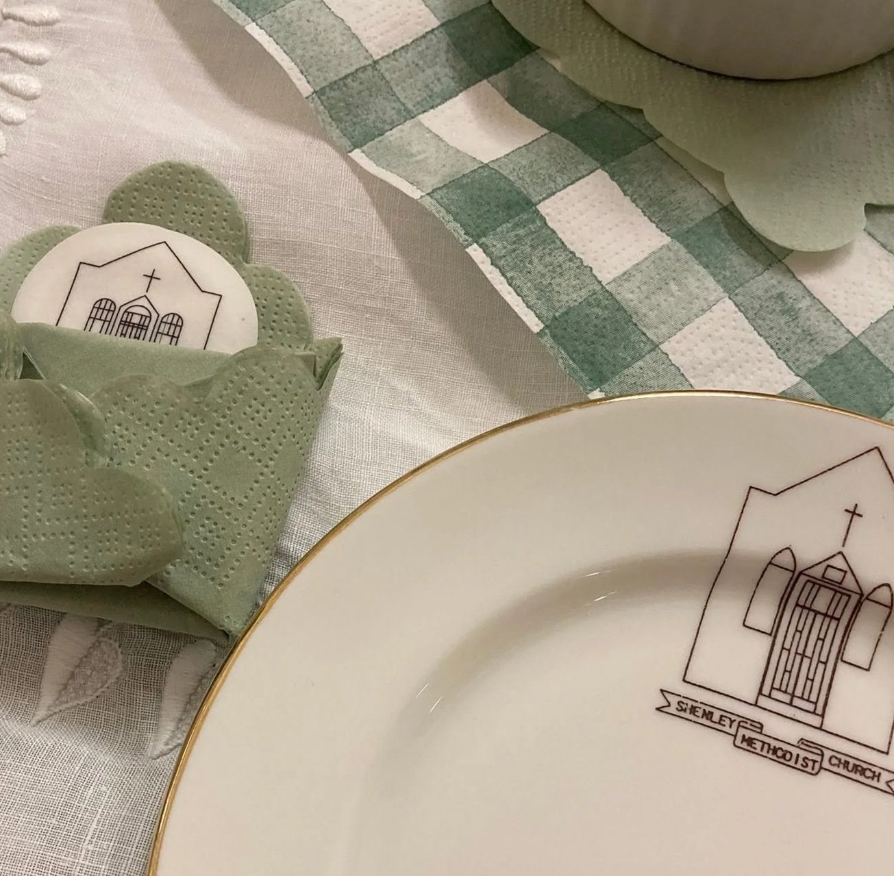 Table setting with a white plate featuring a church logo and gold rim, a green cloth napkin with a church logo, and a green and white checkered table runner.
