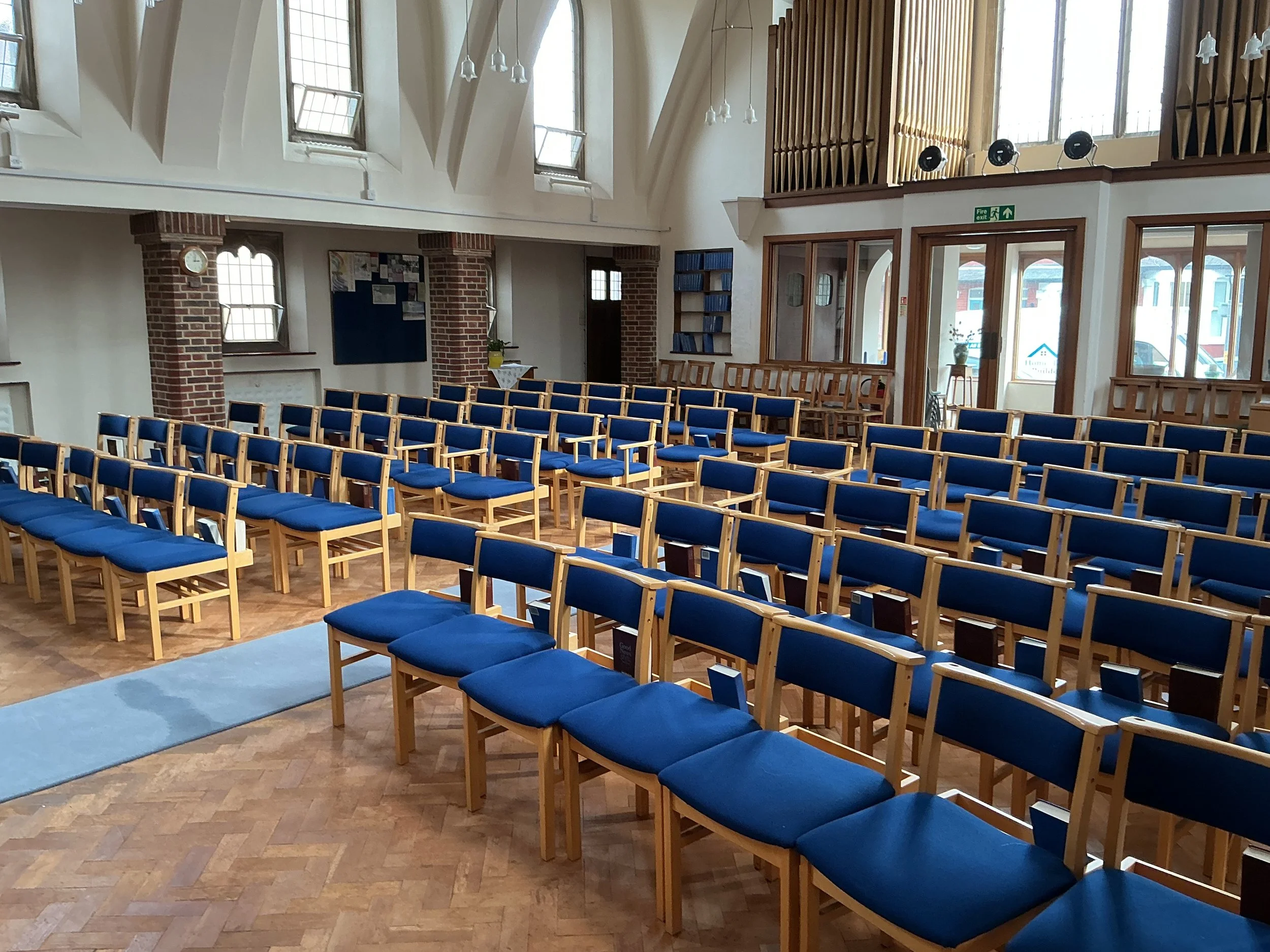 Interior of a church or auditorium with rows of blue cushioned chairs, brick columns, windows, and a balcony with a pipe organ. There are some informational signs and a clock on the wall.