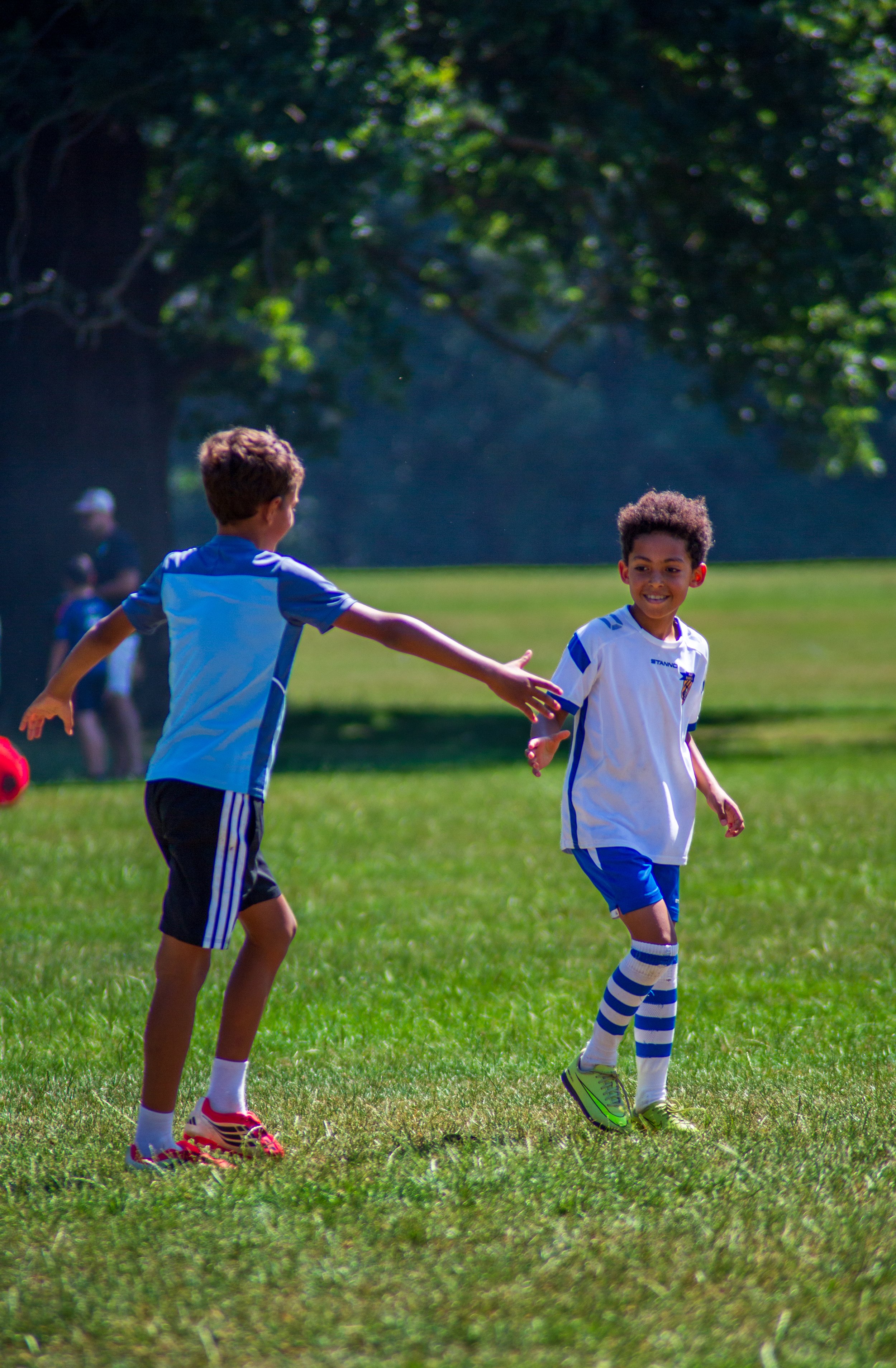 Two young boys playing soccer on a grassy field, smiling and reaching out to each other, surrounded by people and trees in the background.