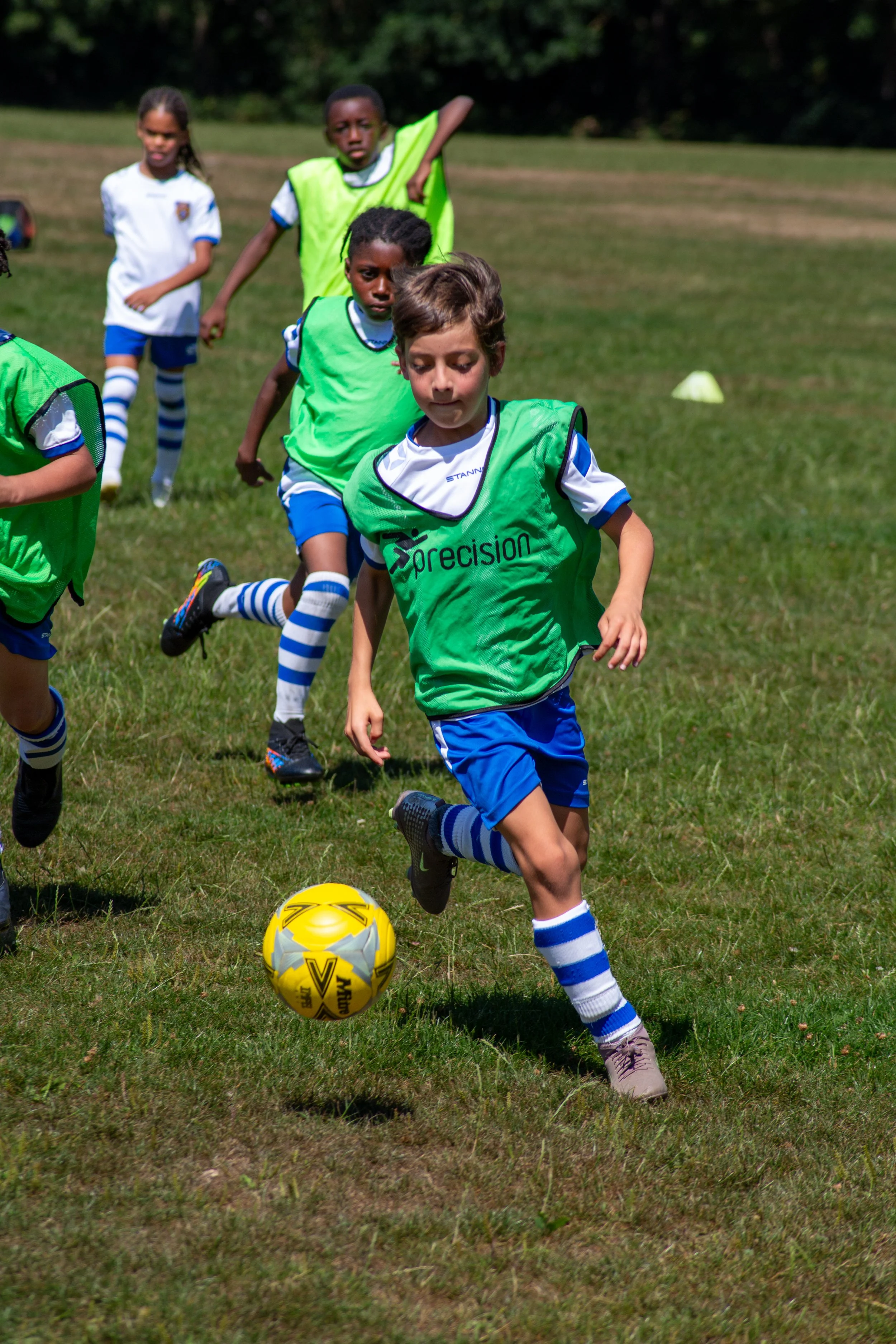 Children playing football on a grassy field, with a yellow football in front of a boy in a green vest and blue shorts, under a clear sky.
