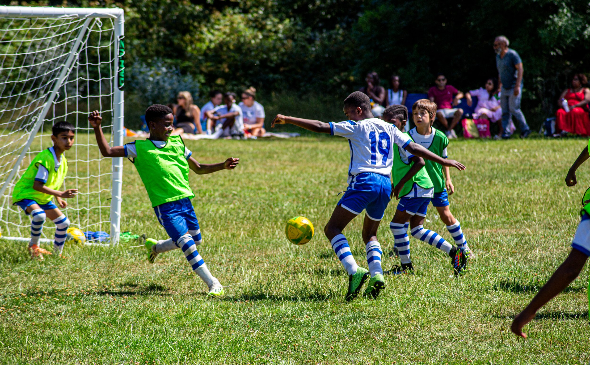 Children playing football on a grassy field during a sunny day, with spectators sitting on benches and standing in the background.