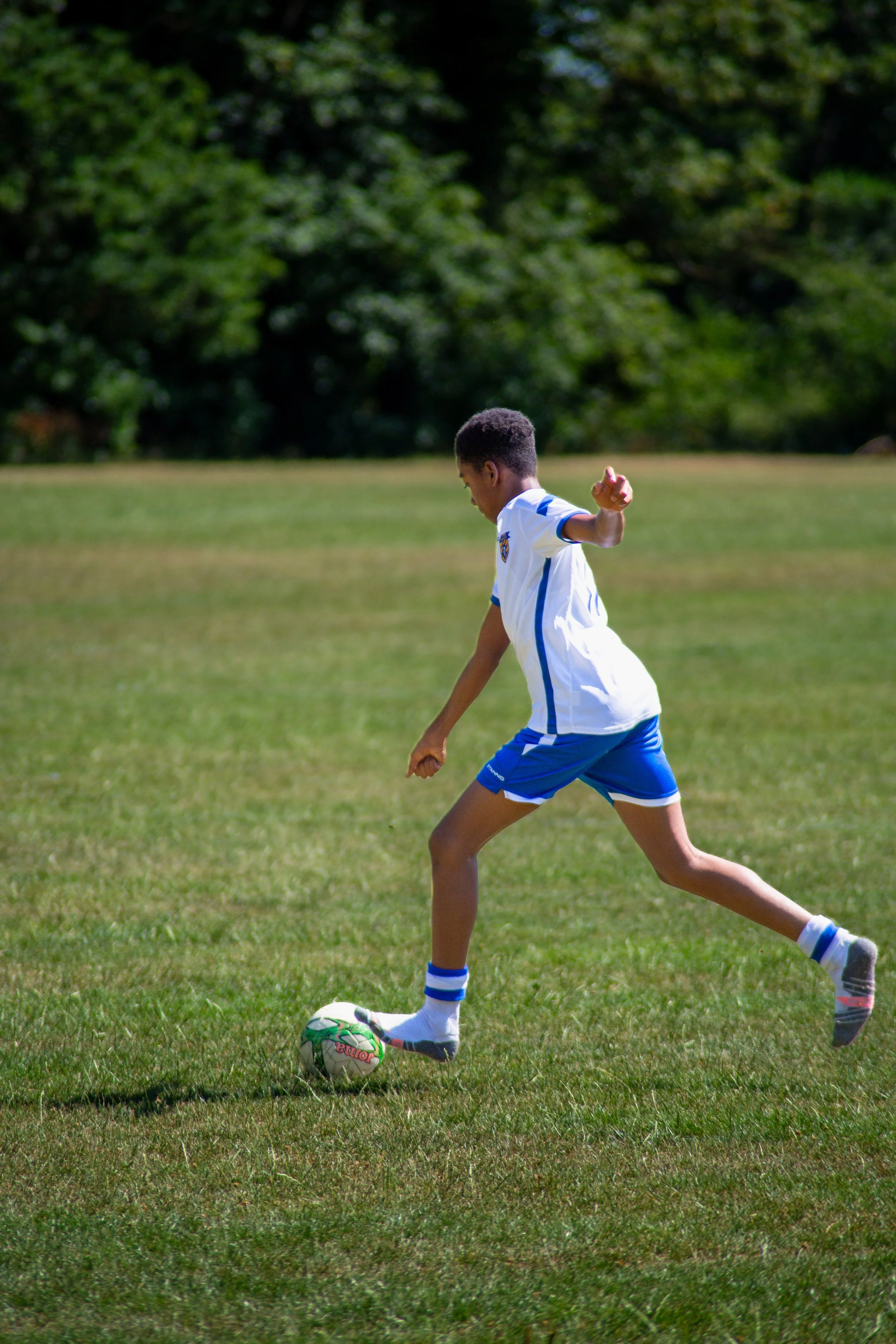 Child in sports uniform kicking a football on a grassy field with trees in the background.