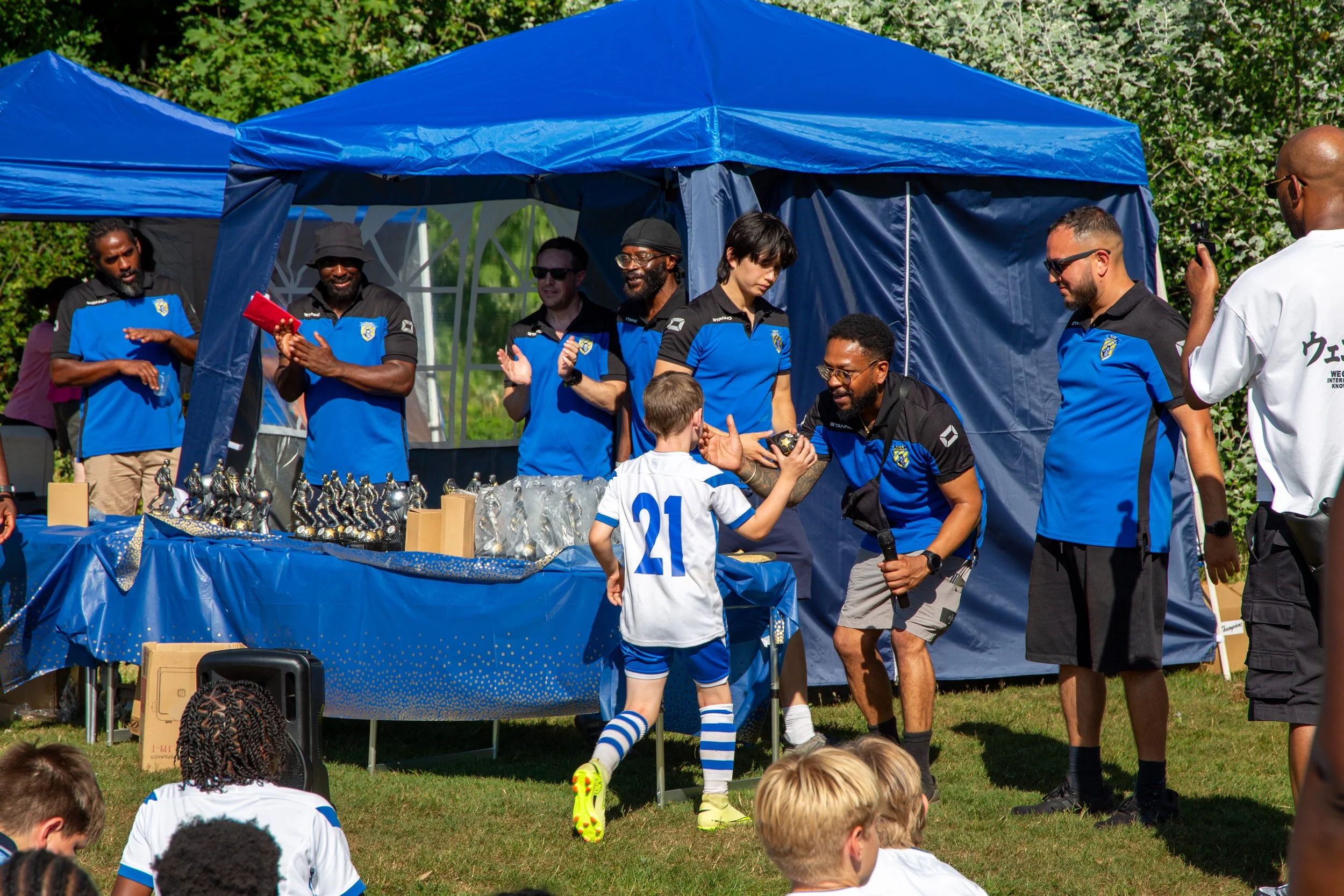 A group of people, including children and adults, gathered at an outdoor event with a blue canopy tent selling sports trophies and awards. Some individuals are wearing black shirts with blue accents, and children are dressed in soccer uniforms. The scene is set in a park with green trees and grass.
