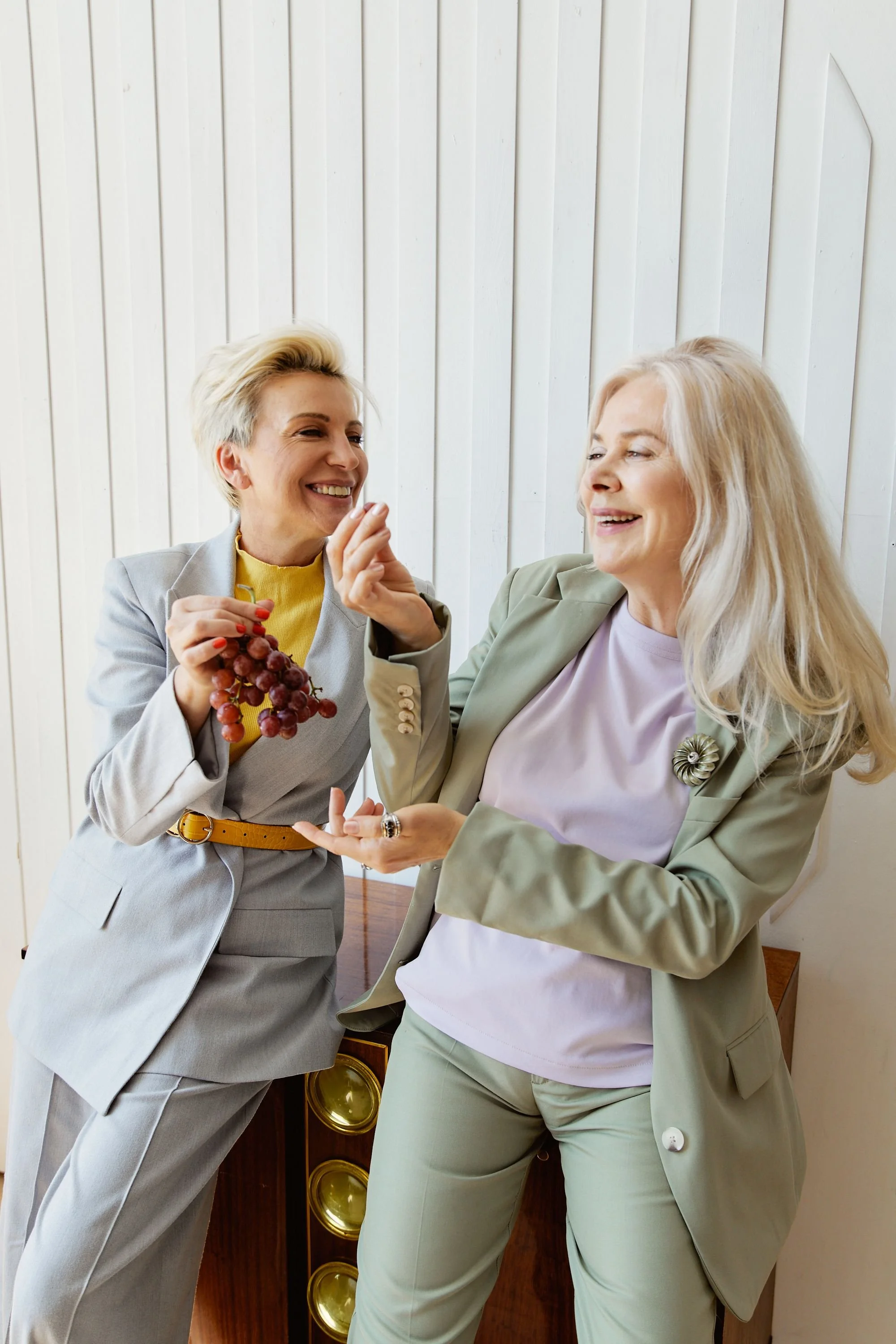 Two women in formal suits sharing a laugh, with one holding a bunch of grapes, in a room with a white wooden wall background.