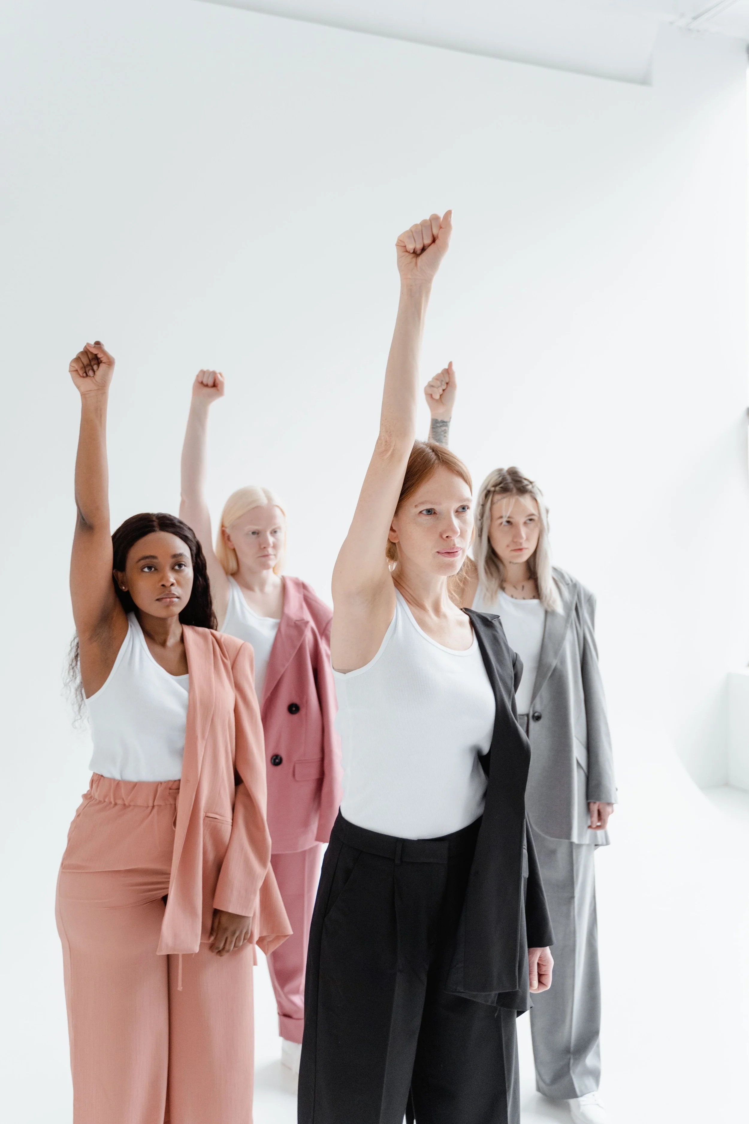 Five women standing in a white room, raising their right fists in the air, wearing business casual and casual clothing in various colors.